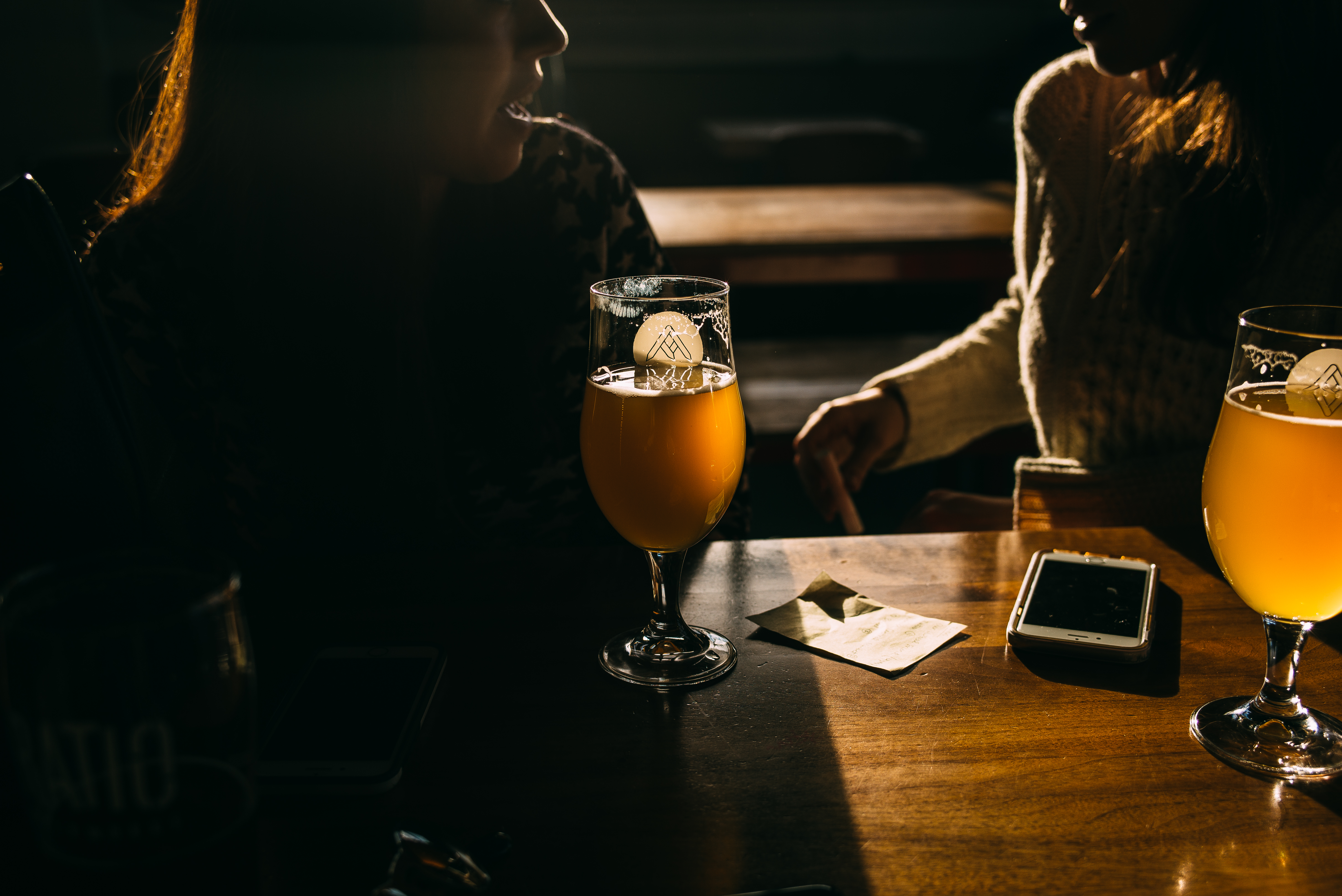 Golden sunlight illuminating two glasses of amber beer on a table with silhouetted patrons behind.