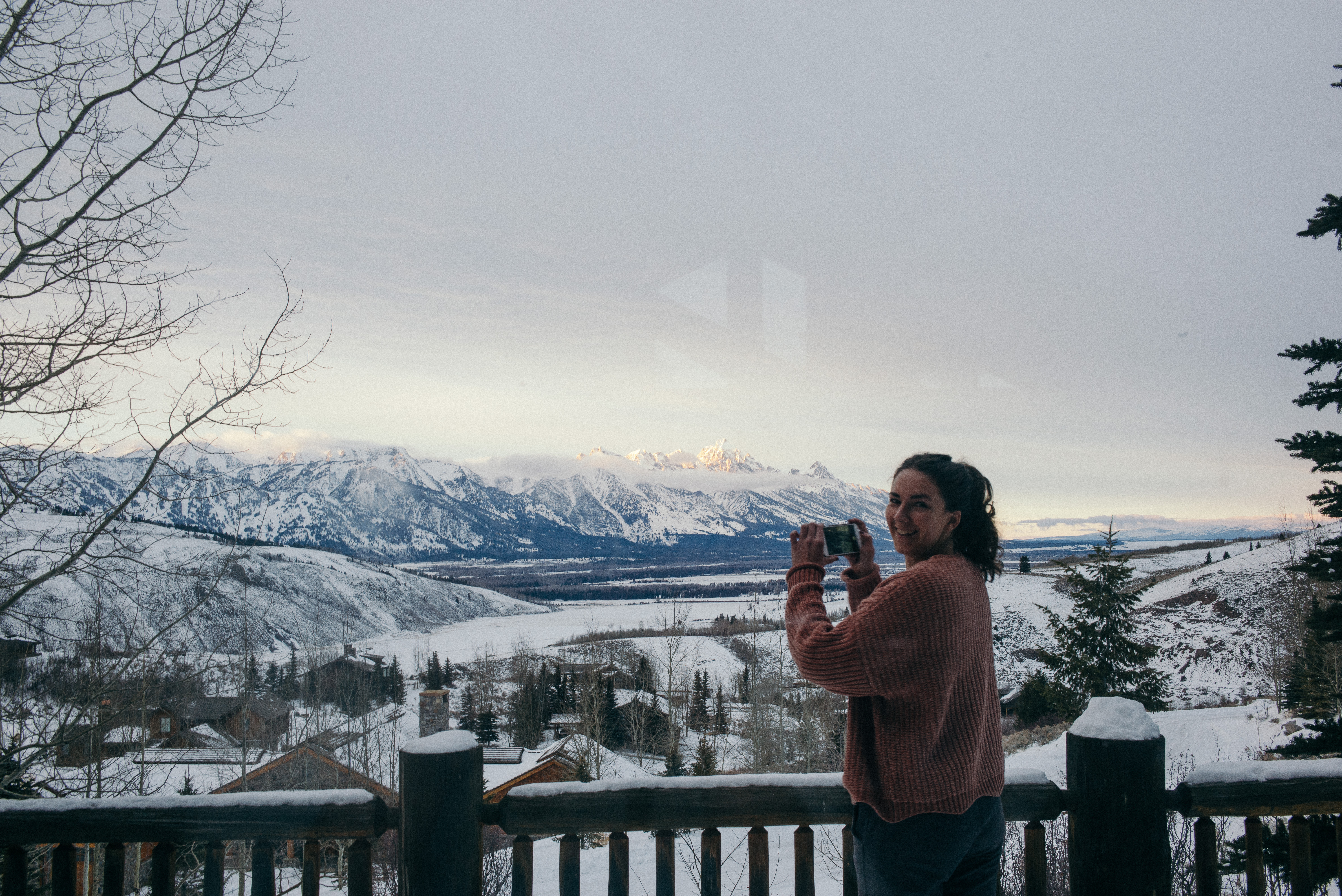 Person in a pink jacket on a snowy balcony photographing the snow-dusted Grand Teton mountain range.