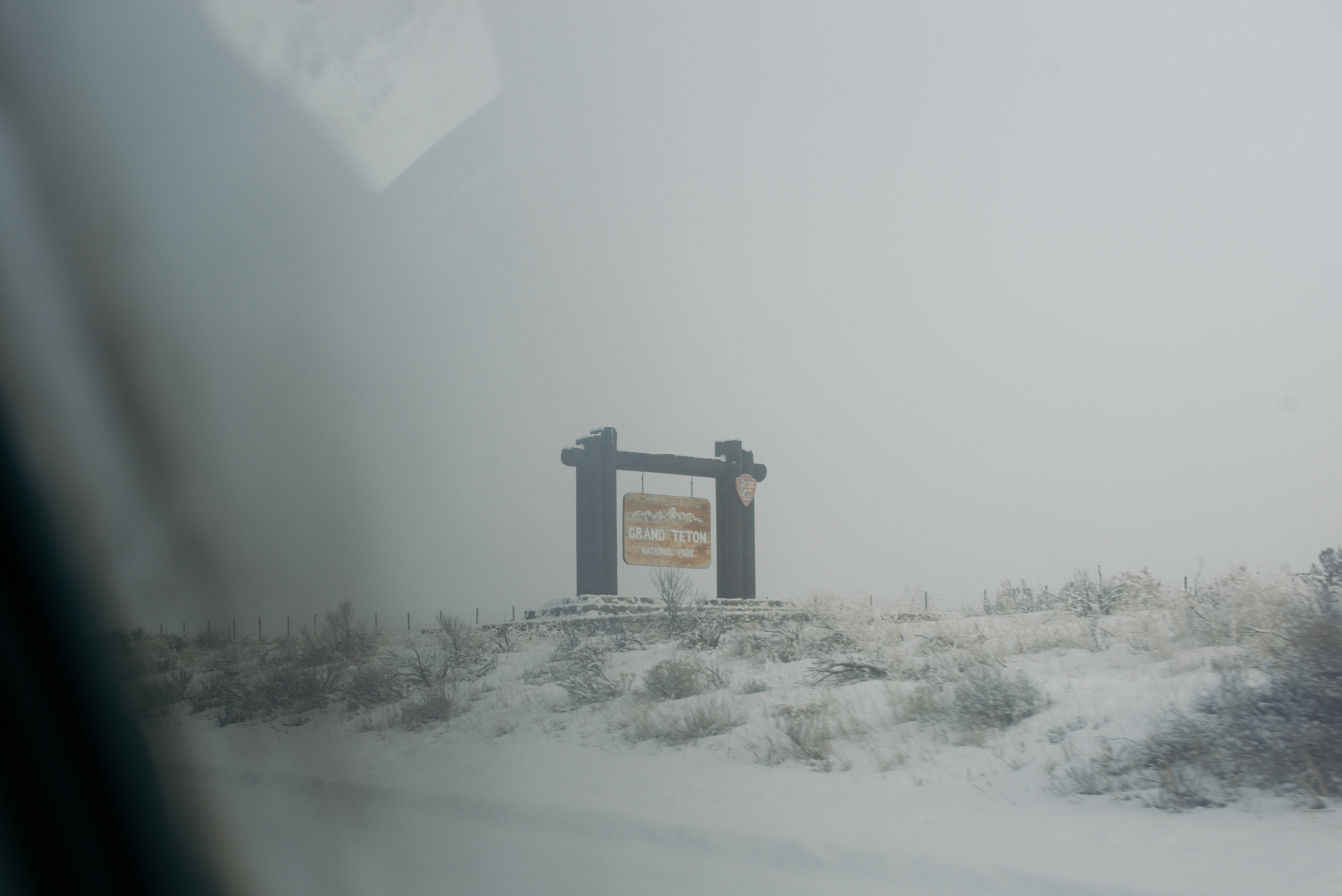 Roadside 'GRAND TETON NATIONAL PARK' entrance sign on a snowy bank amidst thick winter whiteout fog.