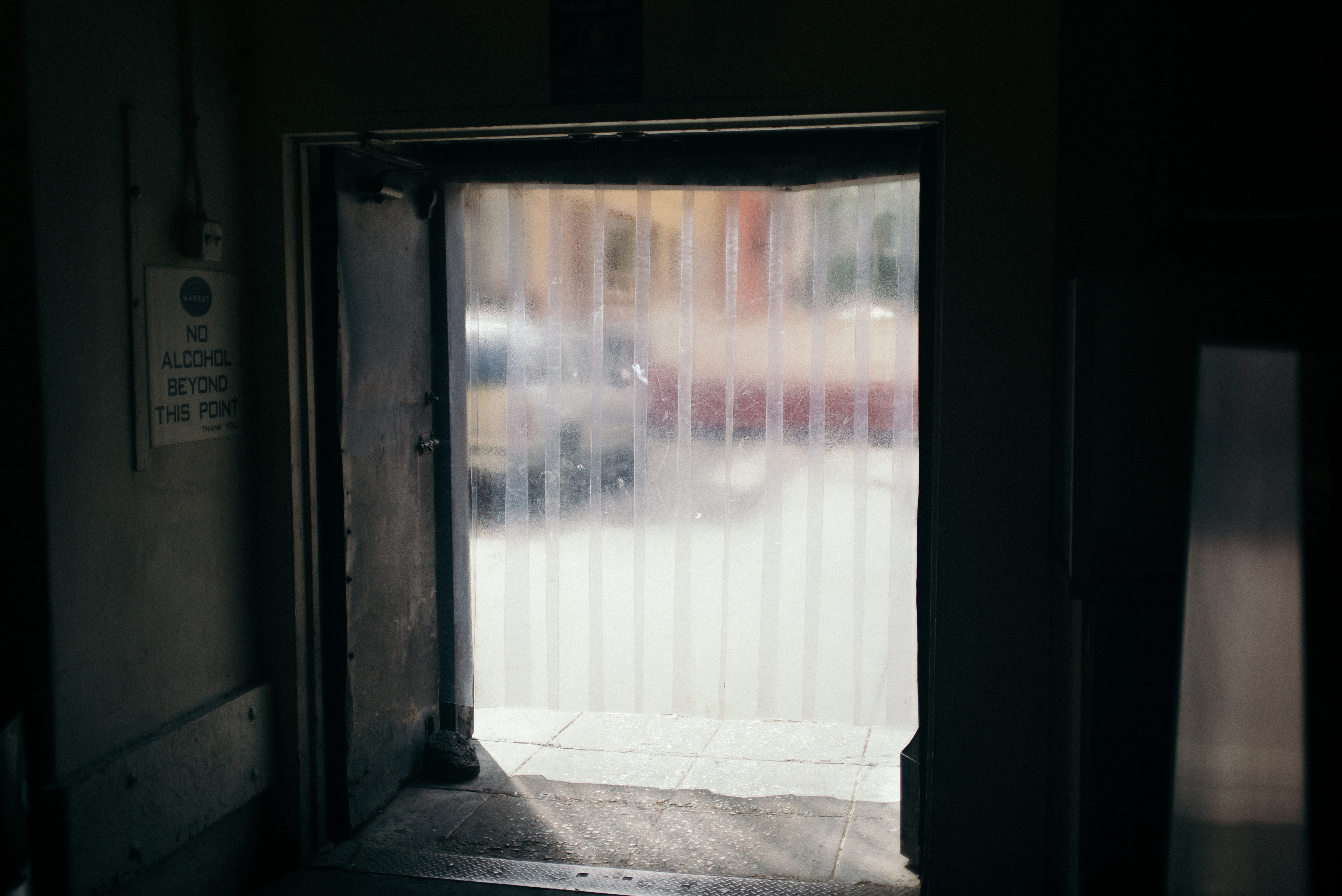 Clear plastic strip curtains looking into an alley beside a 'Market: No alcohol beyond this point' sign.