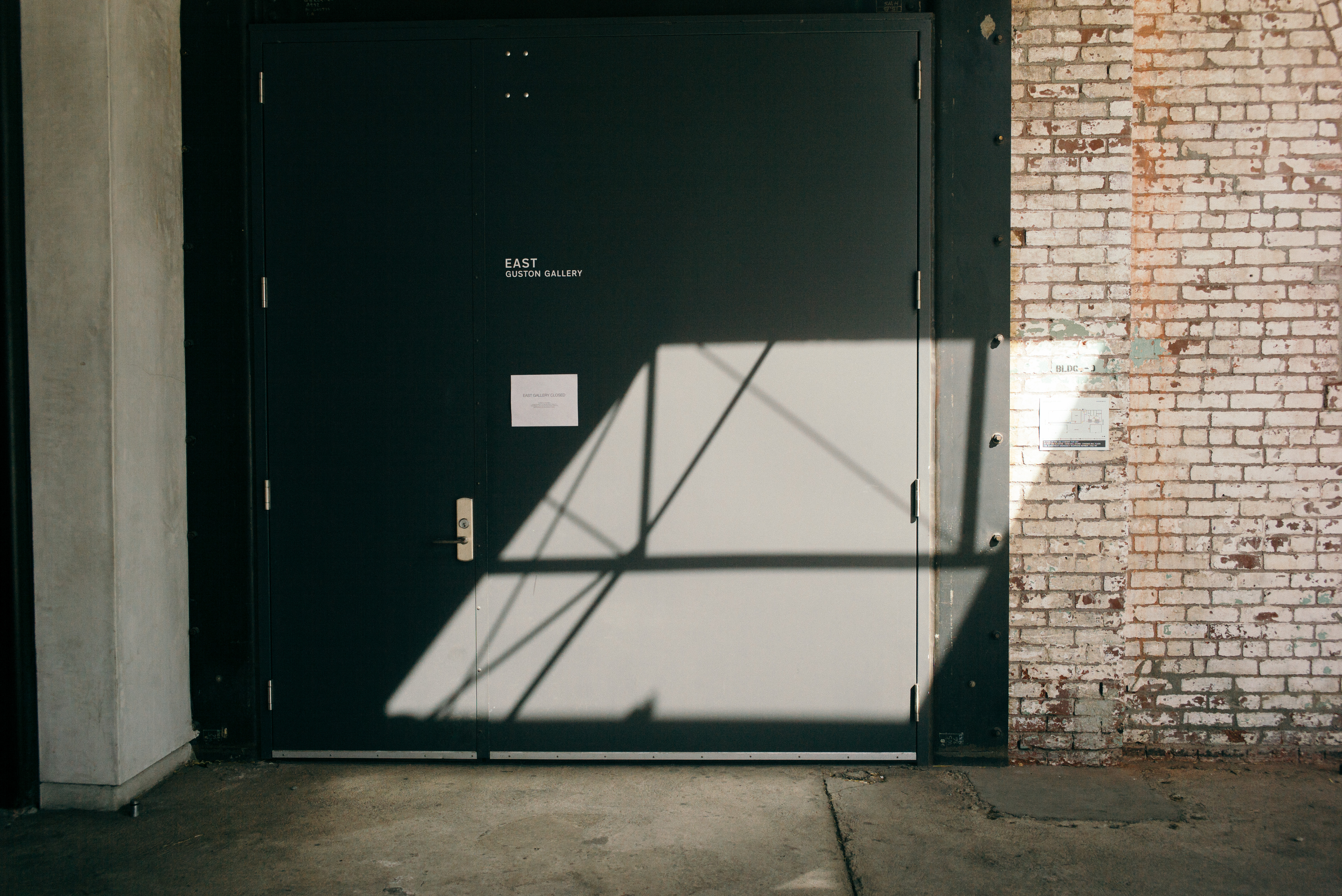 Sunlit white brick facade with dark industrial doors labeled 'EAST GUSTON GALLERY' and geometric shadows.