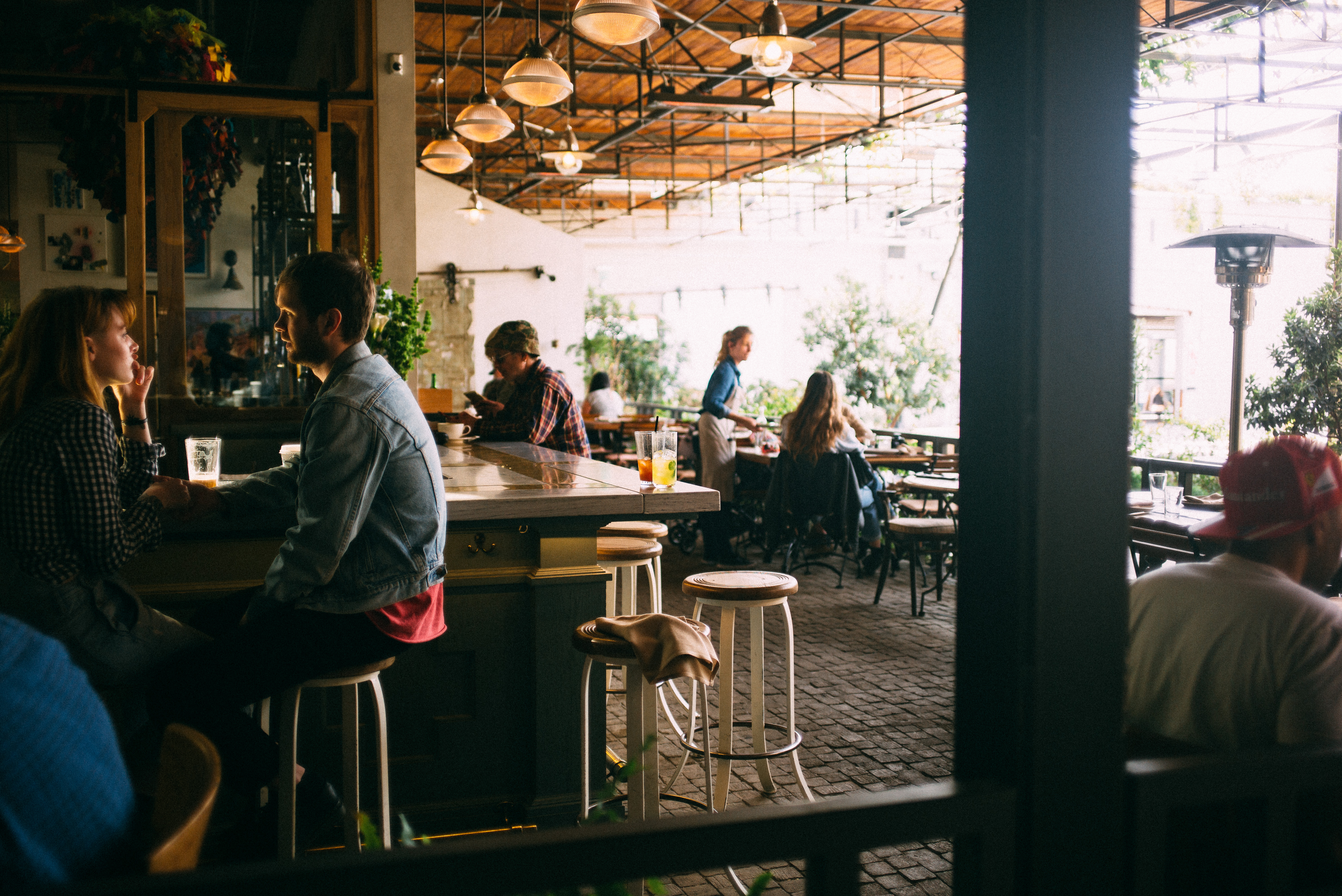 Patrons at an outdoor restaurant bar on a sun-dappled patio with pendant lights and lush potted greenery.
