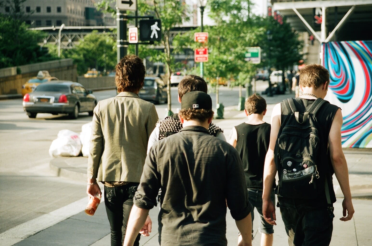 Punk rockers wearing studded vests and backpacks walking down a sunny New York City sidewalk.