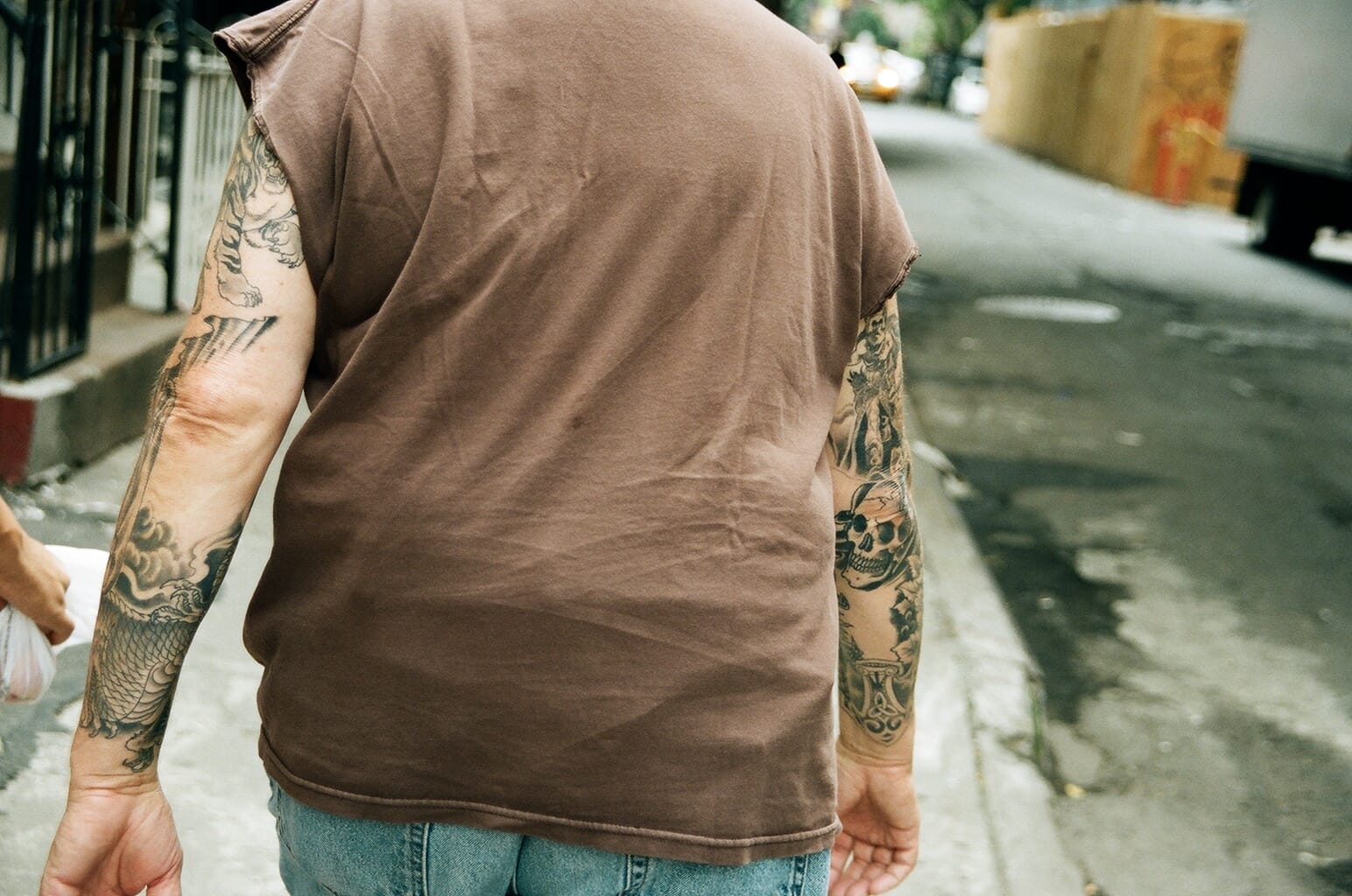 Man walking down a city sidewalk wearing a sleeveless shirt showcasing dragon and skull sleeve tattoos.