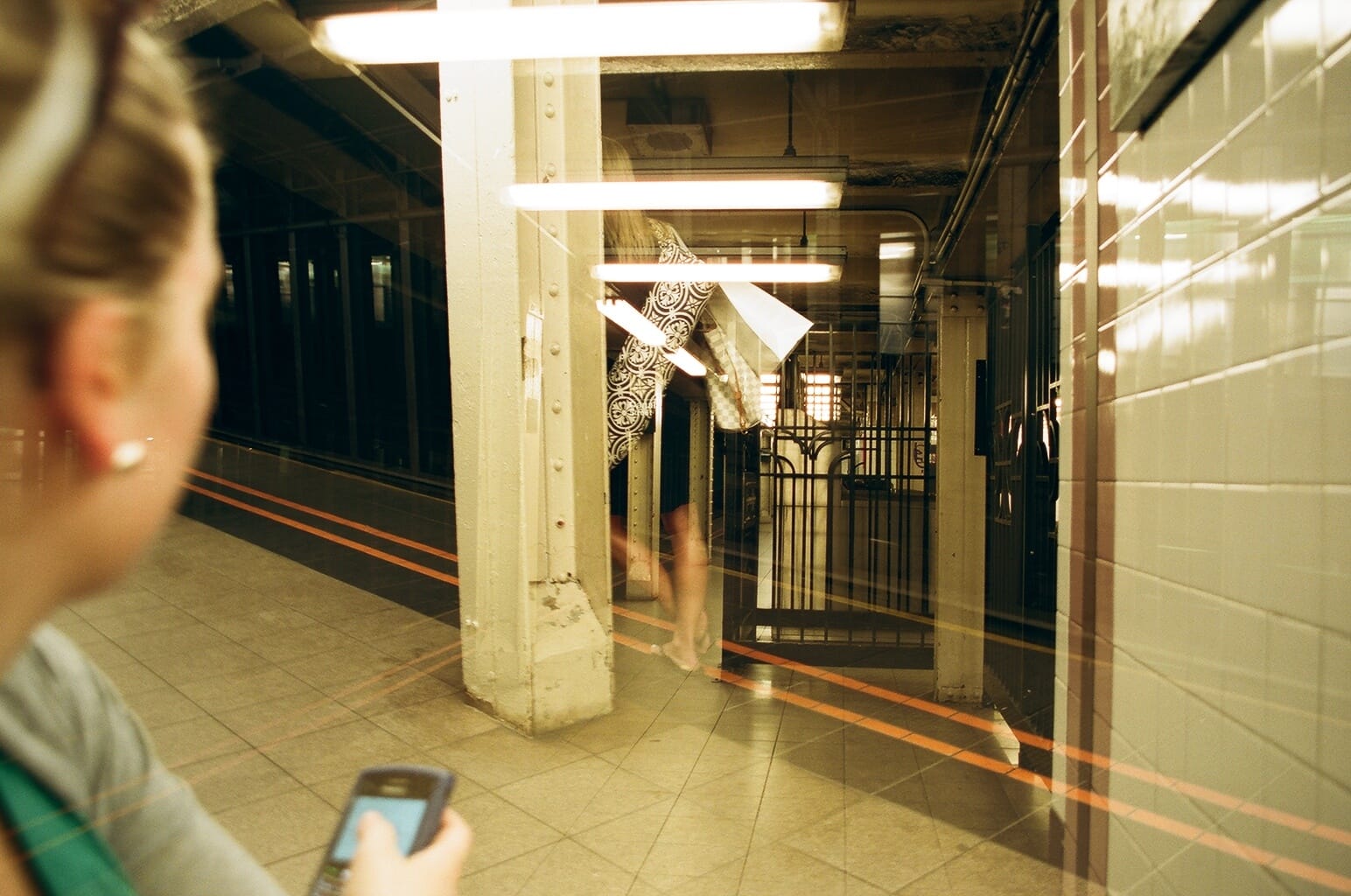 Window reflection from inside a subway car onto an NYC platform as a passenger checks her phone.