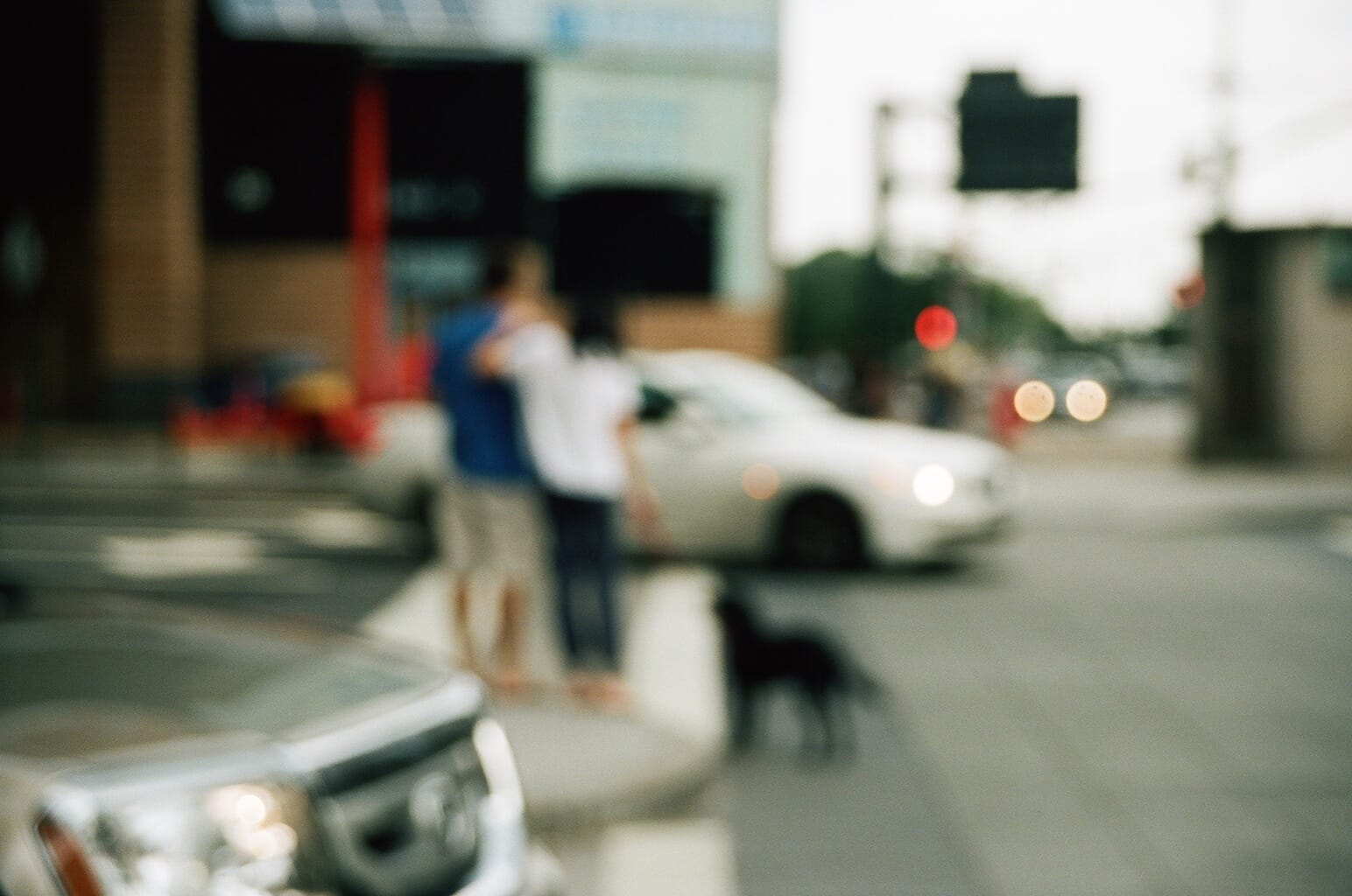 Soft-focus view of a couple with arms around each other walking a black dog across an NYC intersection.