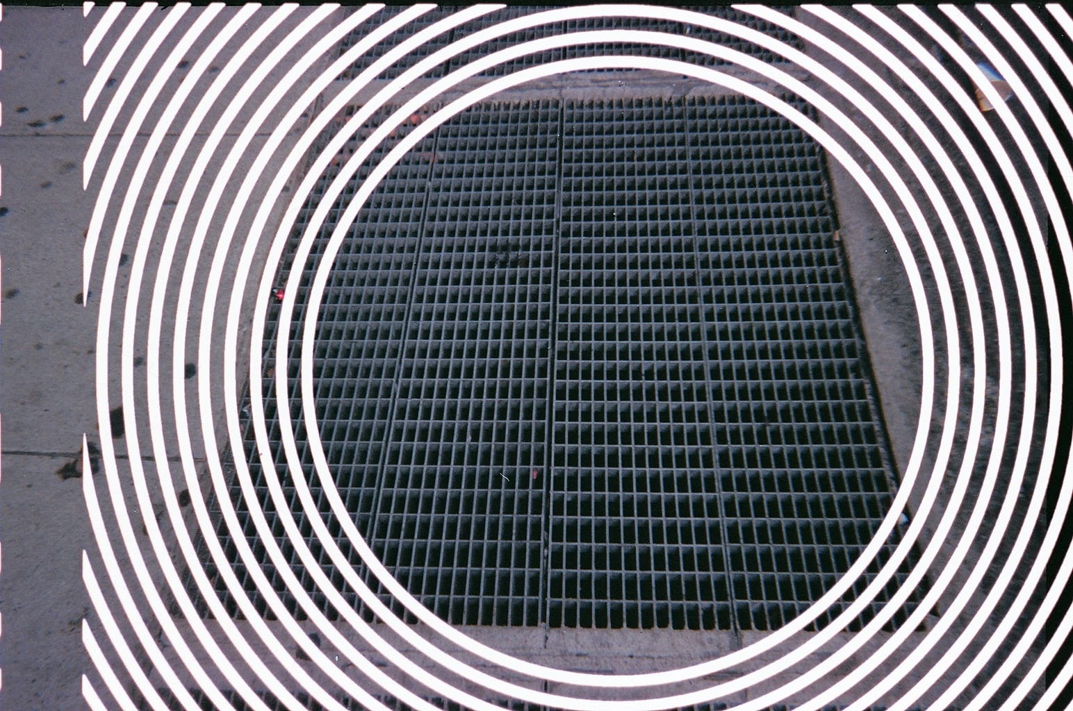 Metal sidewalk subway ventilation grating with white hypnotic concentric rings superimposed.