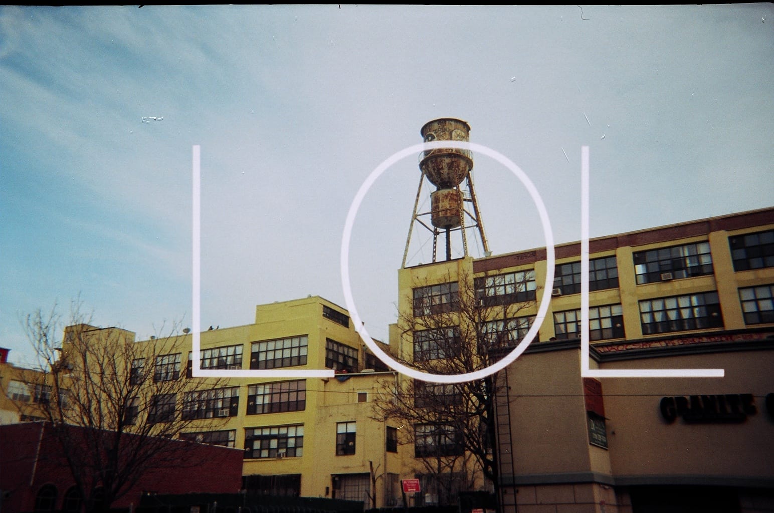 Yellow industrial brick building and rooftop water tower with large white letters spelling 'LOL' superimposed.