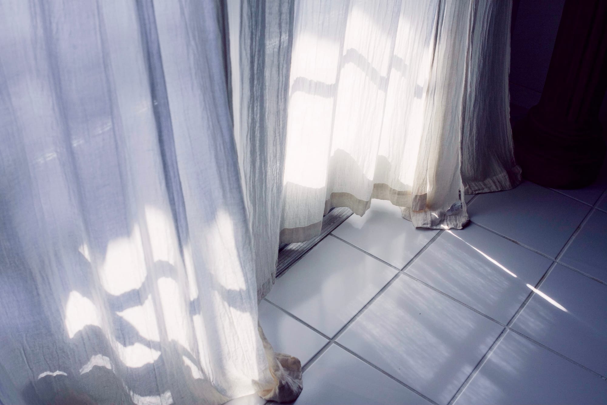 Sunlight through translucent white curtains creating wavy linear shadows on ceramic floor tiles.