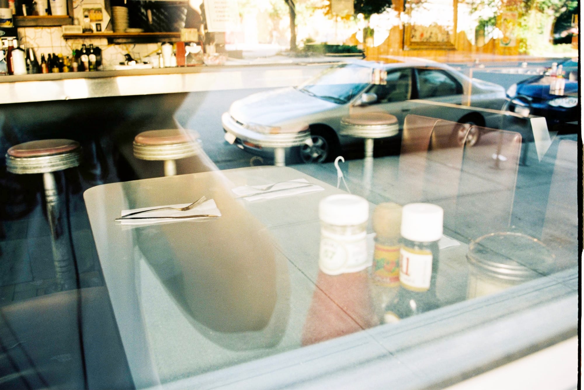 Window reflection in a diner table setting showing condiment bottles, silverware, and a parked sedan.