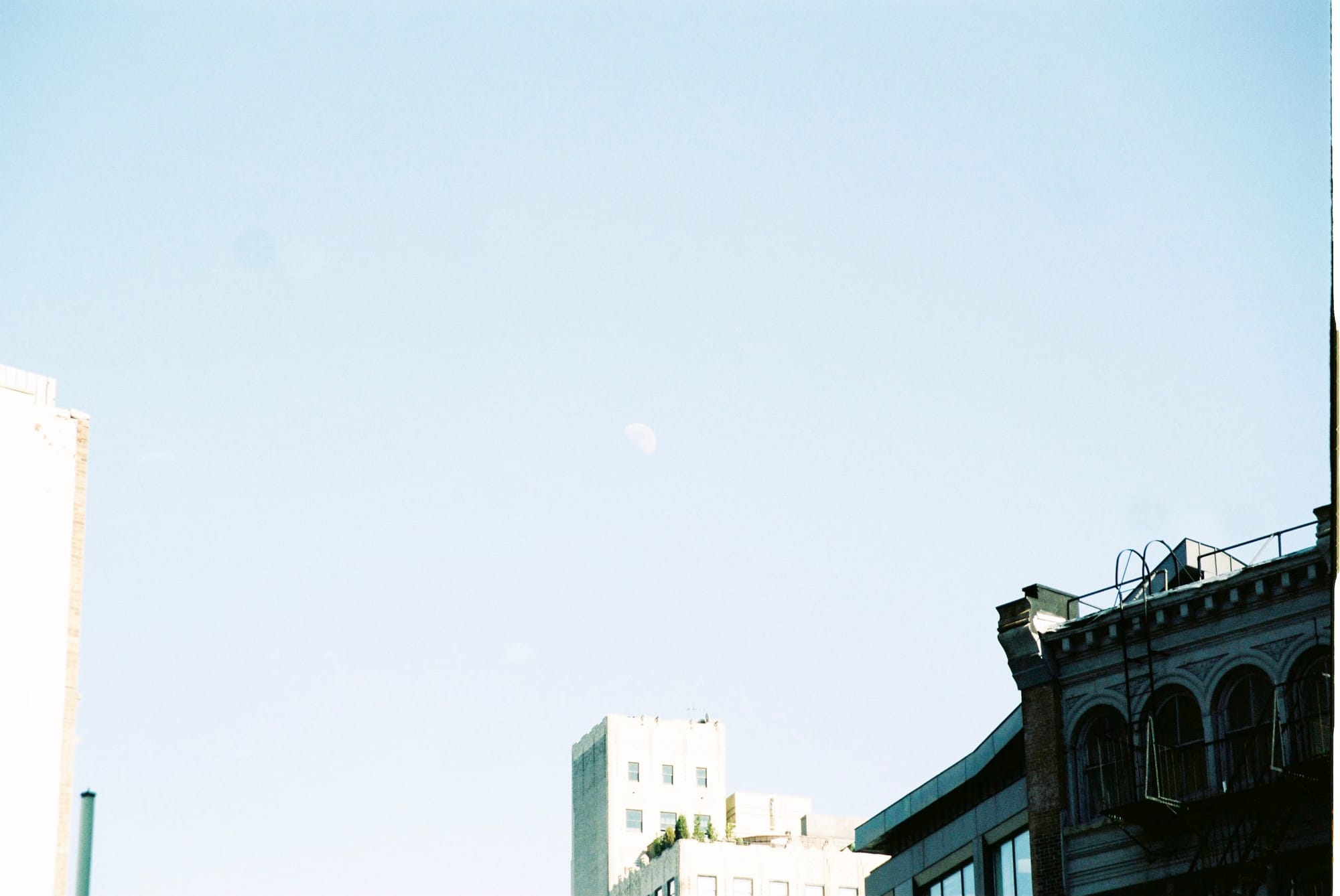 Pale crescent moon hanging in a clear blue daytime sky above historic rooftops in Lower Manhattan.