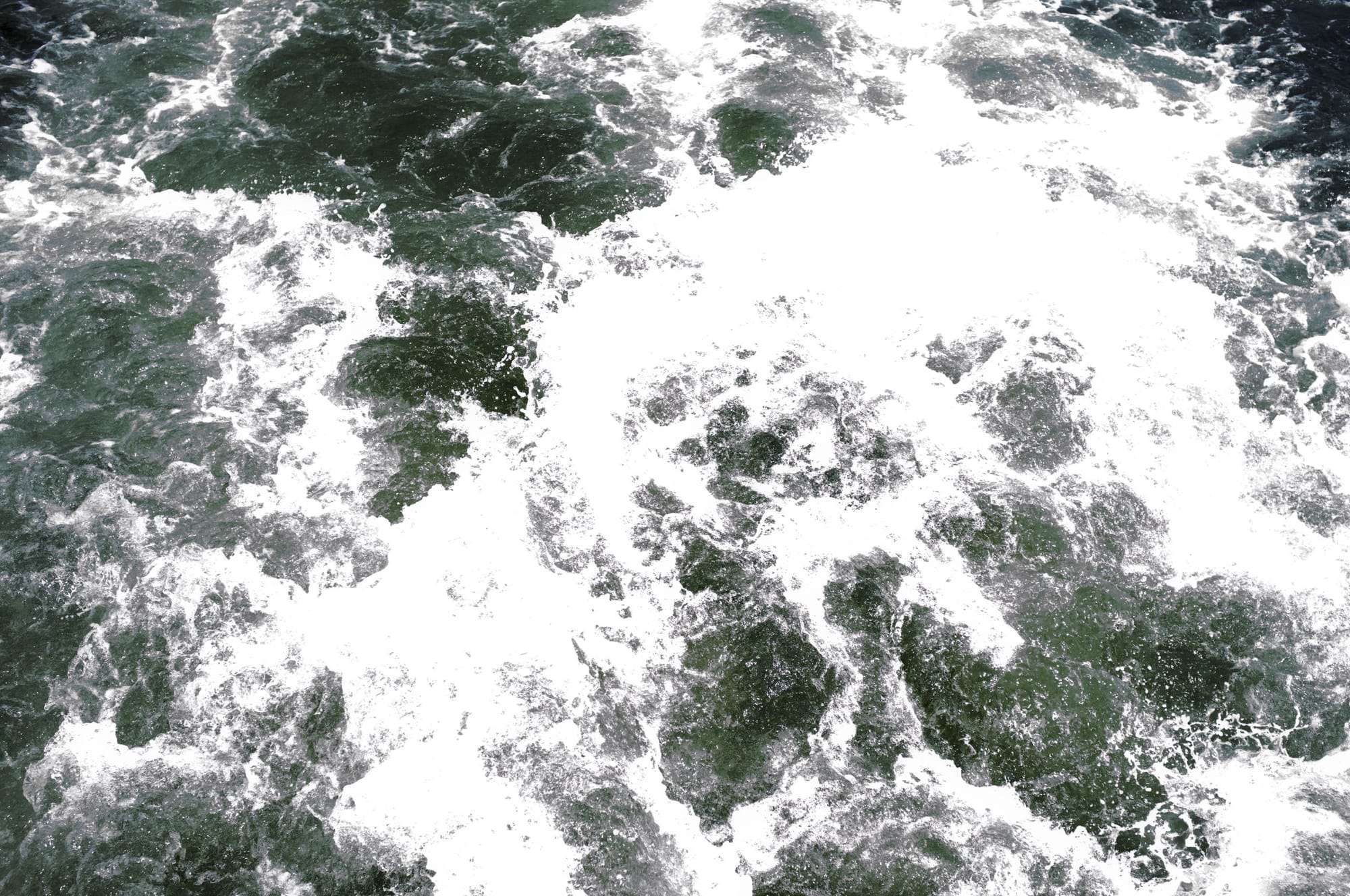 Top-down abstract photograph of churning white foam and deep teal harbor water swirling in the ferry wake.
