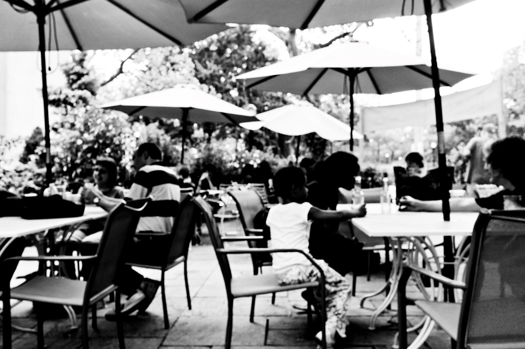 Black and white candid view of families and patrons relaxing under patio umbrellas at the Battery Park Beer Garden.