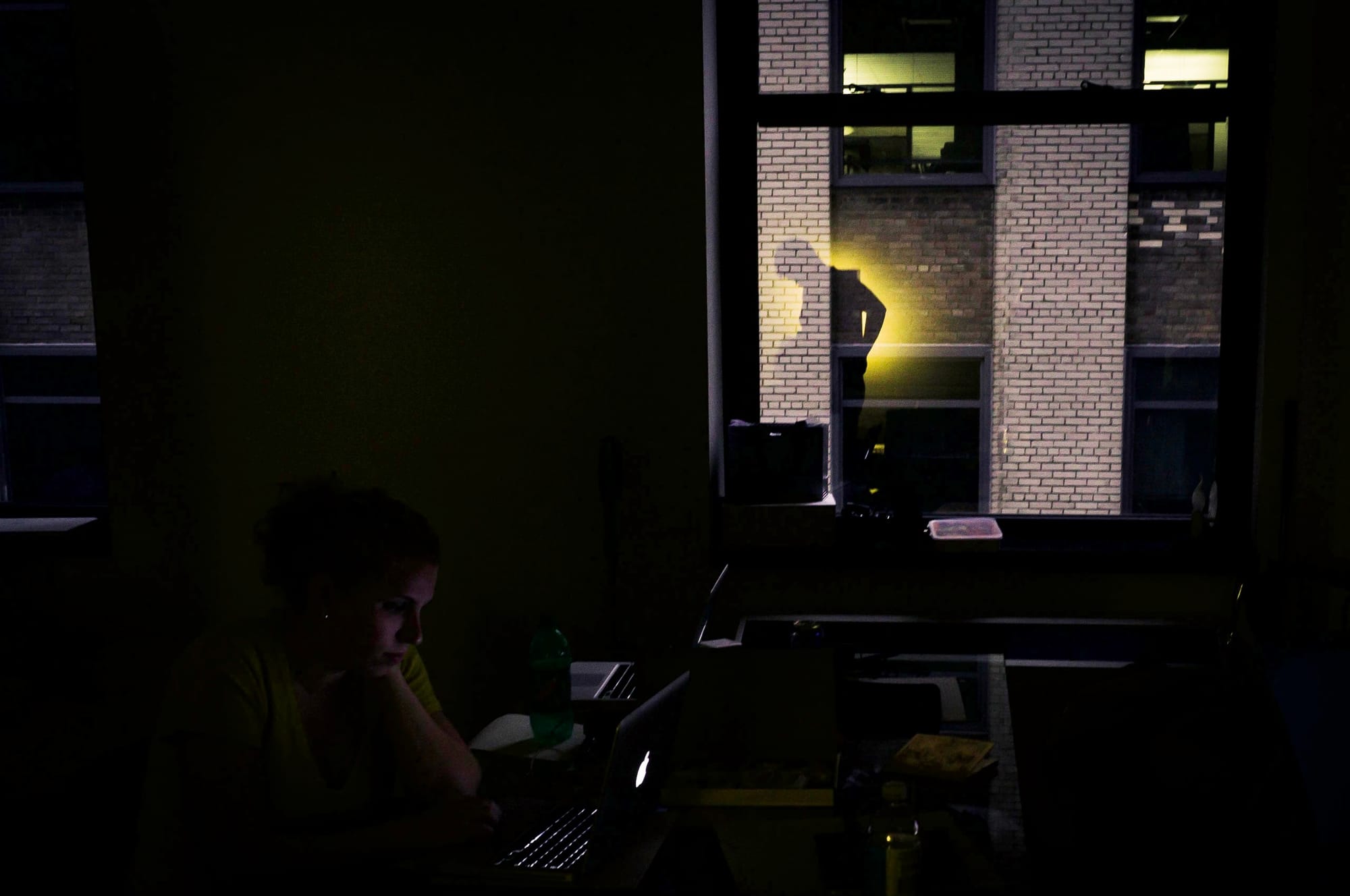 Caitlin working on a MacBook in a dark room with a dramatic shadow cast on the brick wall outside.