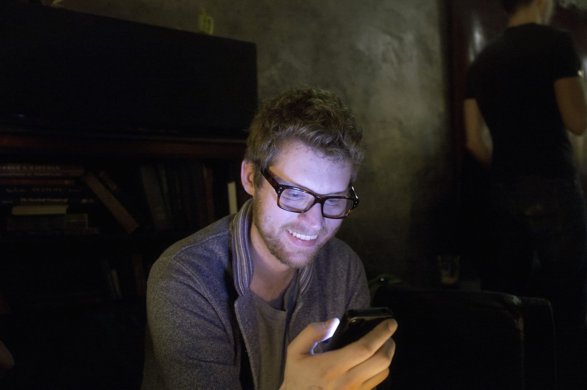 Friend with glasses smiling warmly while looking down at a smartphone in a dim room.