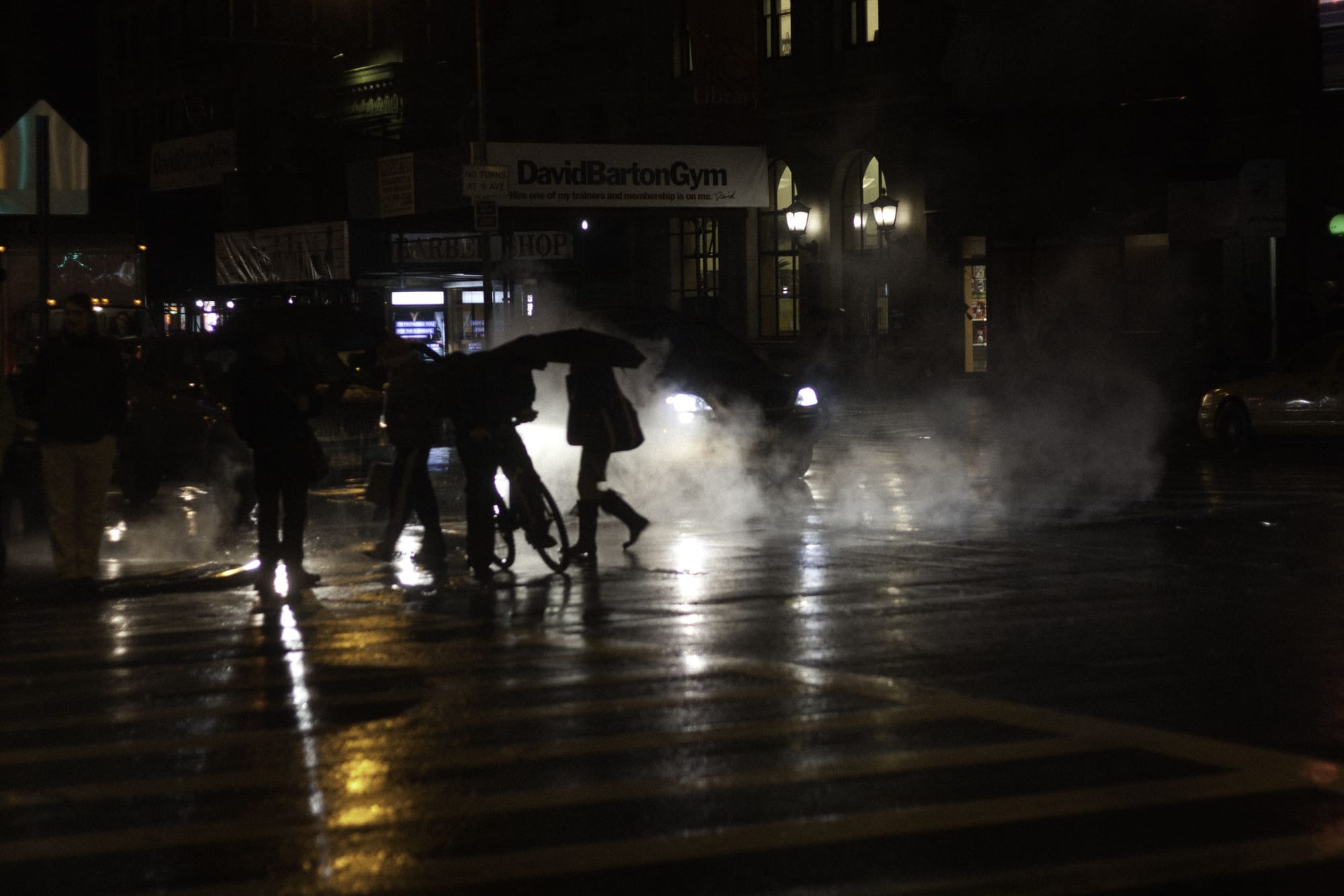 Rain-slicked Manhattan street at night reflecting city lights through rising steam with pedestrians under umbrellas.