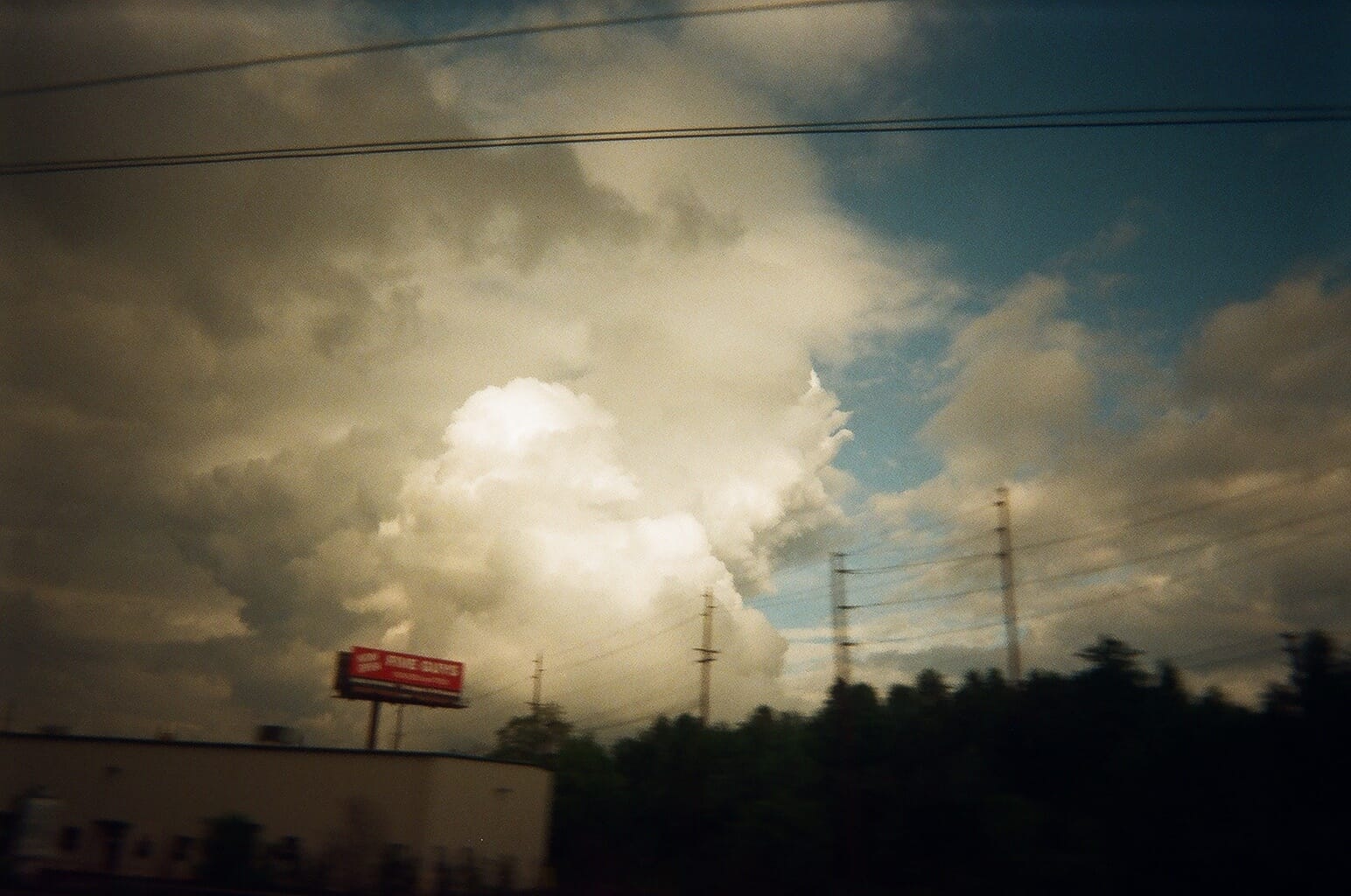 Billowing white cumulus storm clouds towering over a highway billboard and tree line.