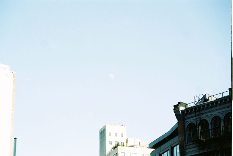 Pale crescent moon hanging in a clear blue daytime sky above historic rooftops in Lower Manhattan.