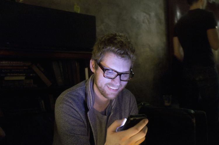 Friend with glasses smiling warmly while looking down at a smartphone in a dim room.