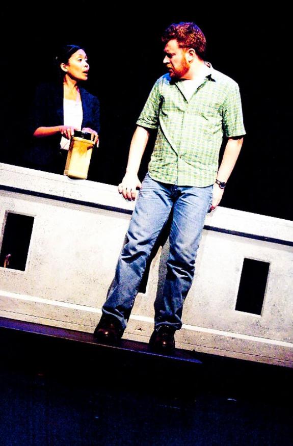 A production show two actors on stage. The characters are talking while one is standing outside a bridge railing while the other stands on the bridge