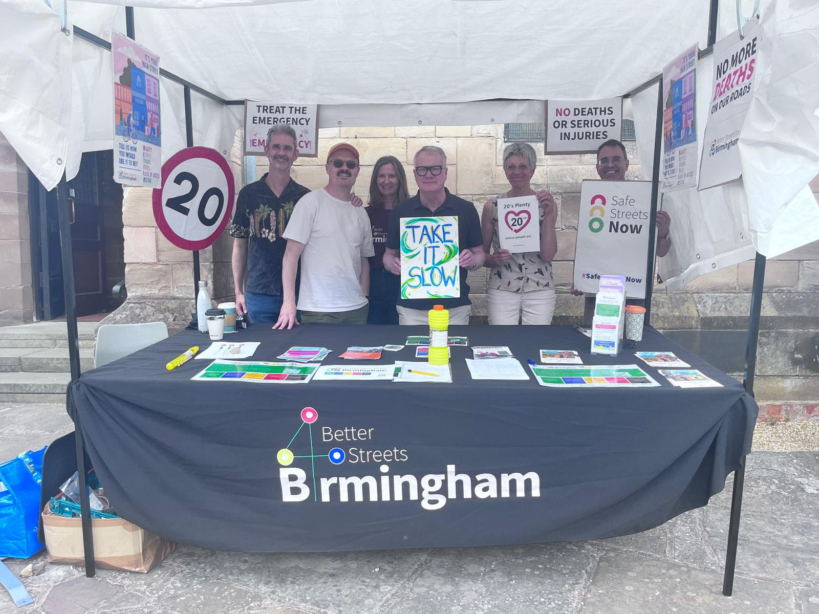 A Better Streets for Birmingham stall, covered in posters and leaflets about the road safety emergency. There are six people stood behind the stall. Left to right: Gareth Hooper, Rob Kewley, Kirsten de Vos, West Midlands Mayor Richard Parker holding a sign that reads "Take it slow", Councillor Izzy Knowles holding a sign that reads "20's Plenty" and Councillor Tix Huxtable holding a sign that reads "Safe Streets Now".