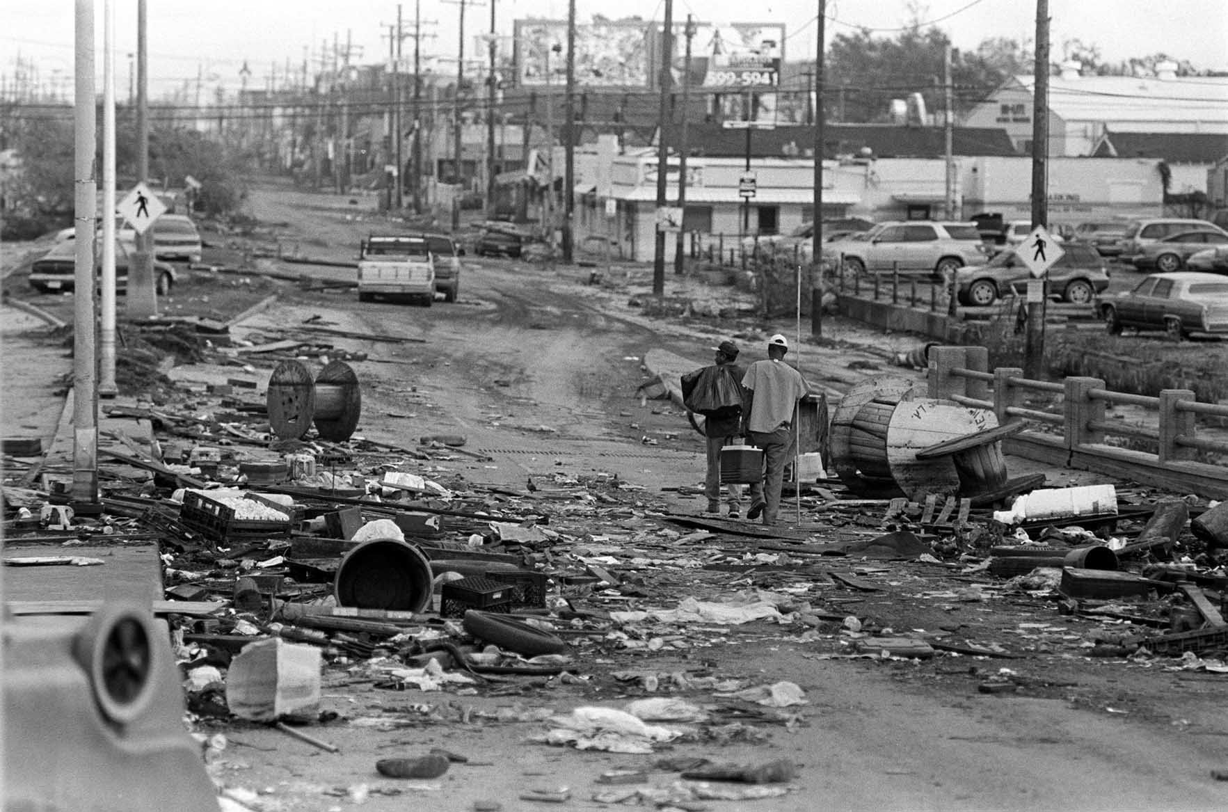 "Prison Debris." New Orleans, 2005. Inkjet Print on Canvas. Photographed in the days after Hurricane Katrina, this image depicts two men walking through debris-strewn streets as the city struggled to recover. To their left, out of frame, looms the old Orleans Parish Prison, where the incarcerated were abandoned as floodwaters engulfed the facility. Their presence near the facility evokes both the vulnerability and resilience of New Orleanians, standing as testimony to survival, neglect and the enduring fight for dignity in the face of disaster. Credit: Clarence Williams