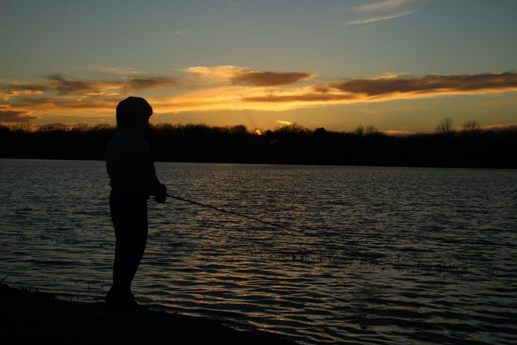 Young boy holding fishing pole over body of water with trees and sunrise/sunset in the background.