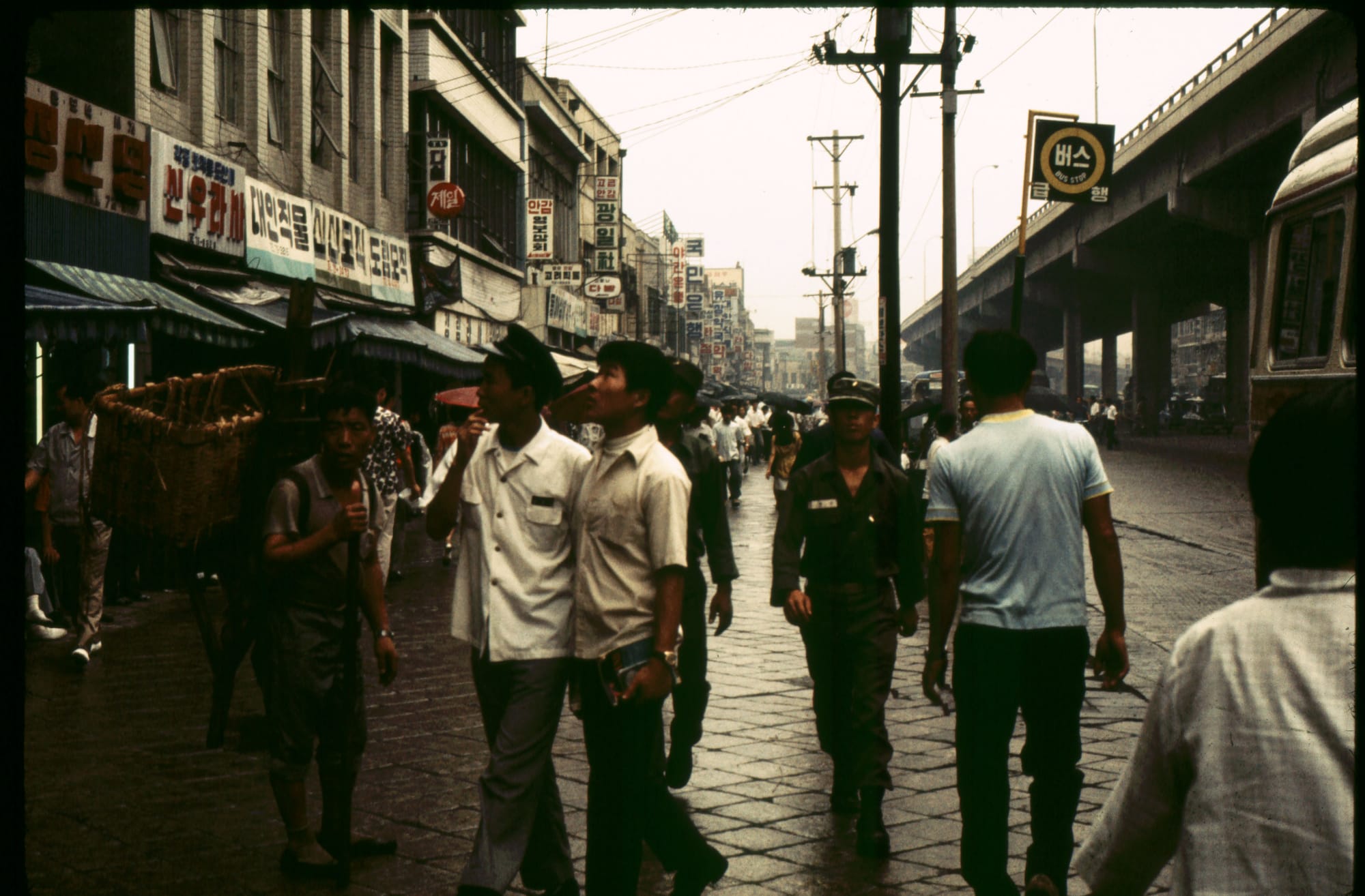 1970 年首爾雨天街景,街道兩側可見韓文招牌、行人與公車