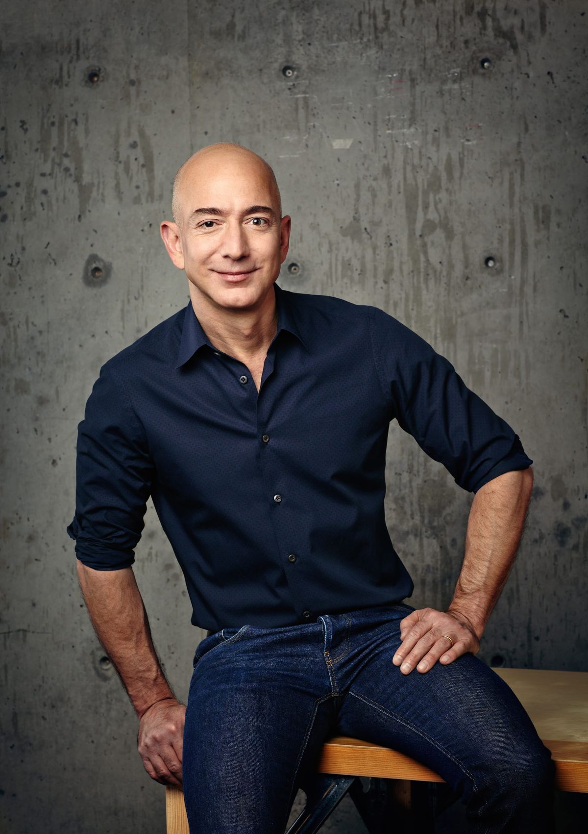 Amazon 官方媒體庫提供的 Jeff Bezos 肖像，穿深藍色襯衫坐在灰色牆前