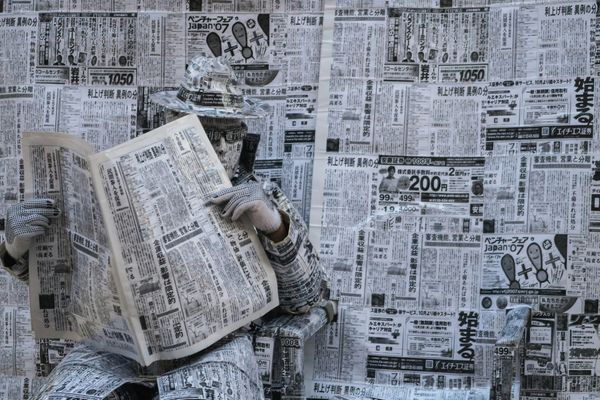 A man wrapped in newspaper from head to toe, reading a newspaper in front of more newspapers.