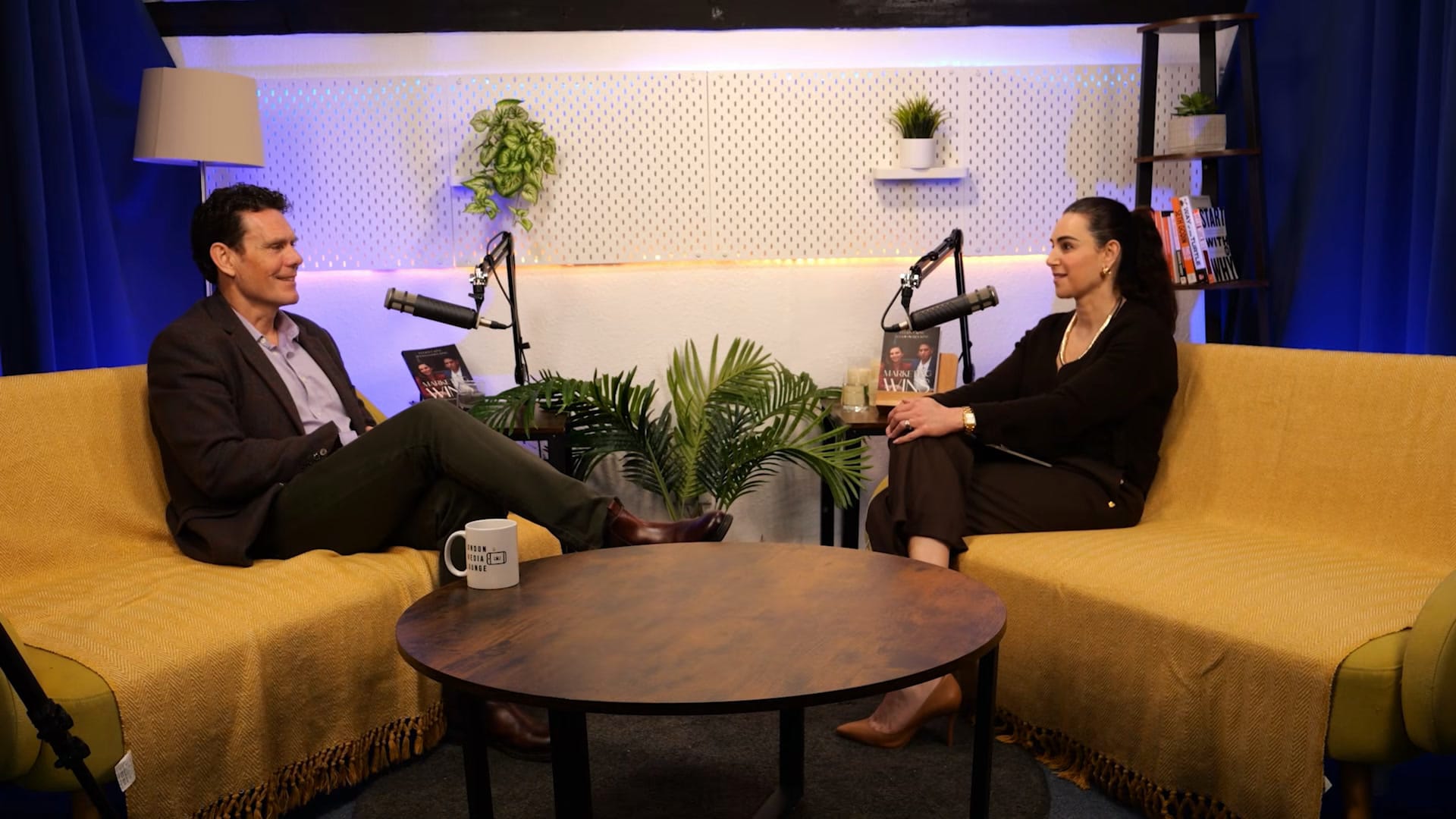 Steve Featherstone and Aleksandra King facing each other across a round table on the BEYOND set, both on mustard sofas with boom microphones between them
