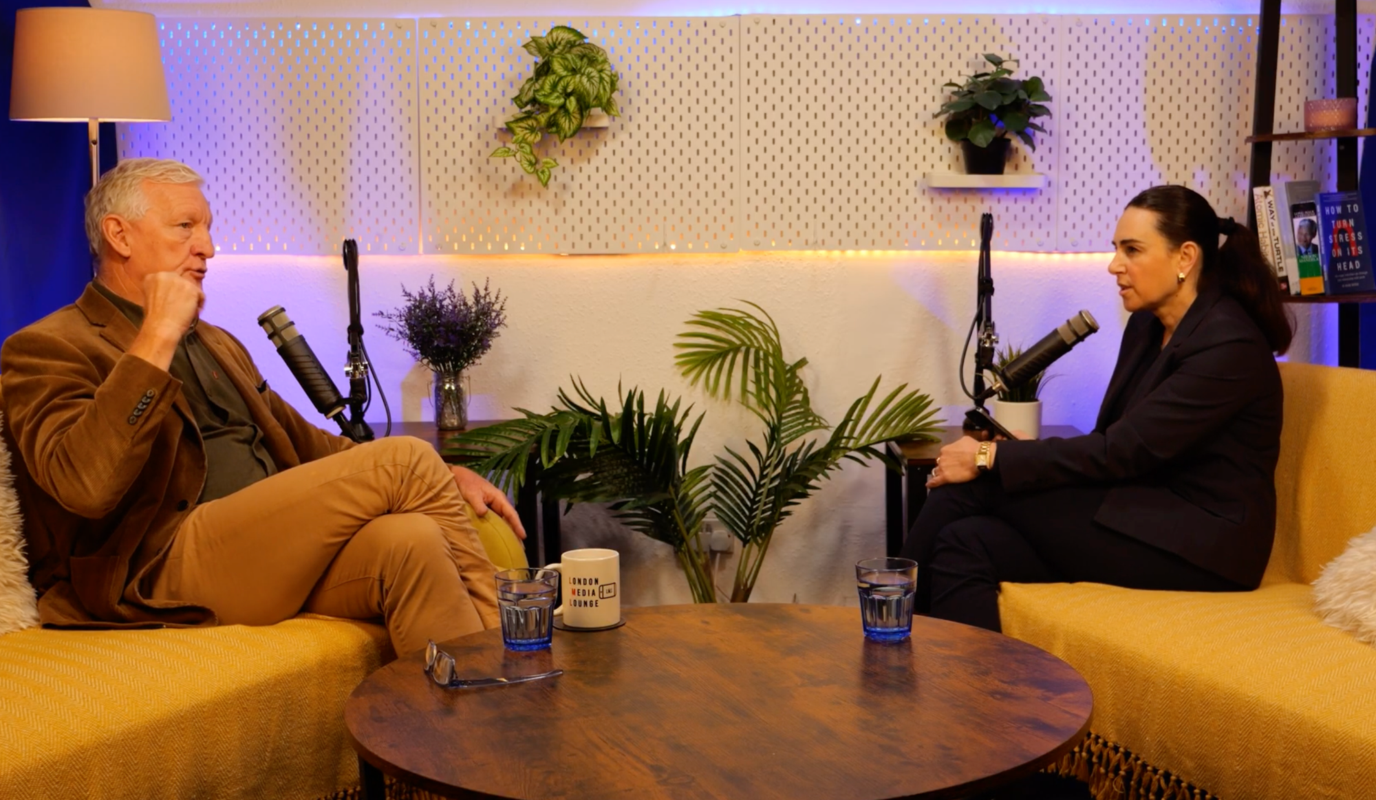 Derrick McManus in a tan corduroy jacket and host Aleksandra King in conversation on the BEYOND set, seated on yellow sofas either side of a round wooden table