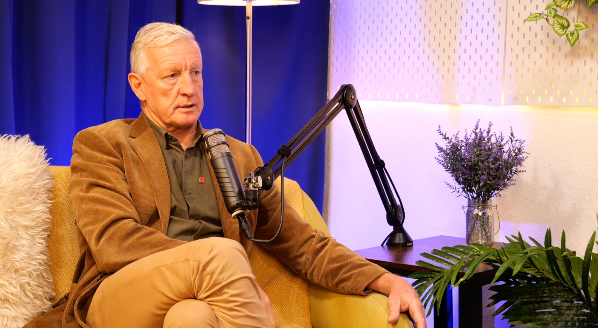 Derrick McManus speaking at the microphone during his BEYOND interview, in a tan corduroy jacket with a poppy pin