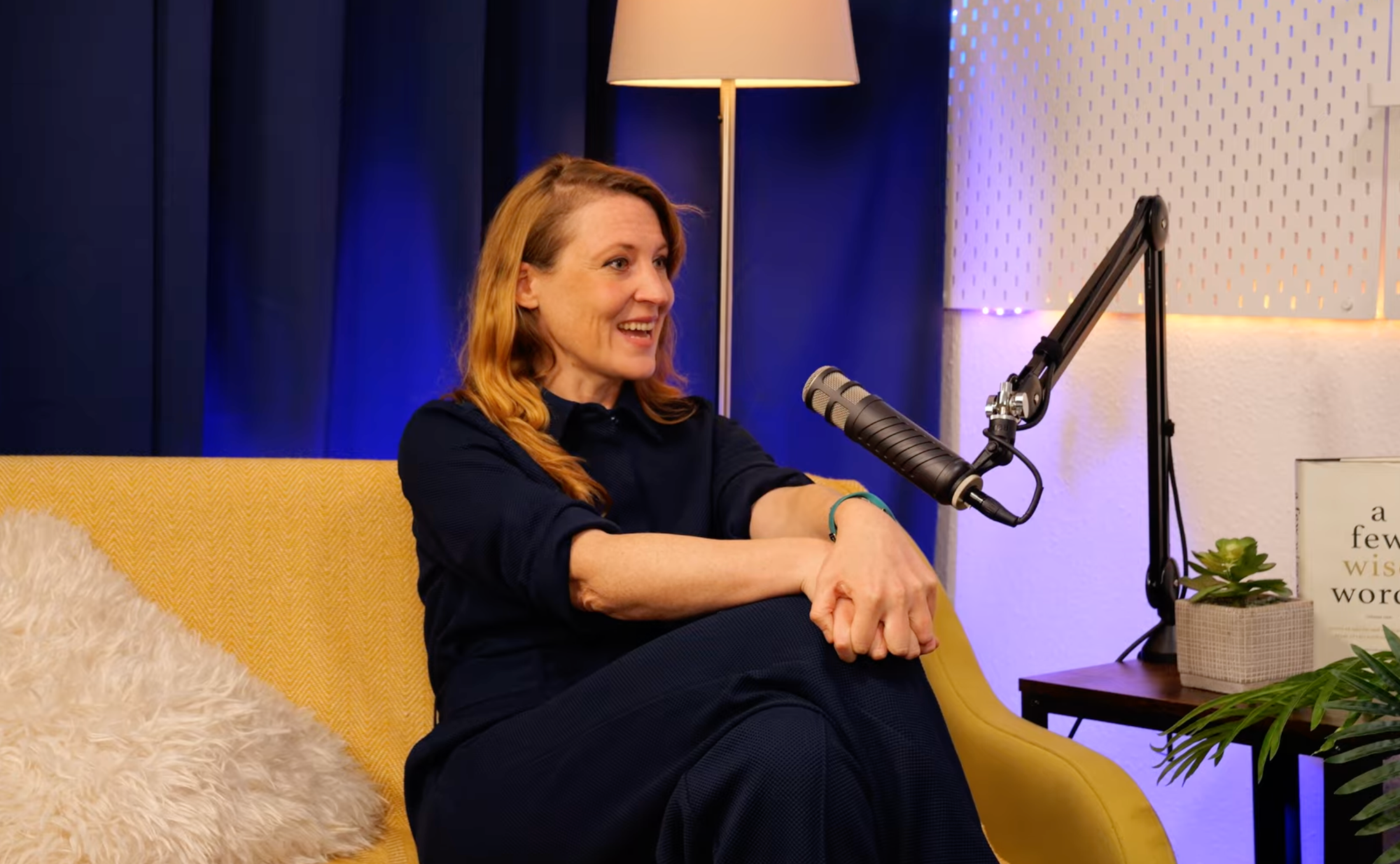 Emily Hughes smiling mid-conversation on the BEYOND set, seated on a yellow sofa with a copy of A Few Wise Words beside her
