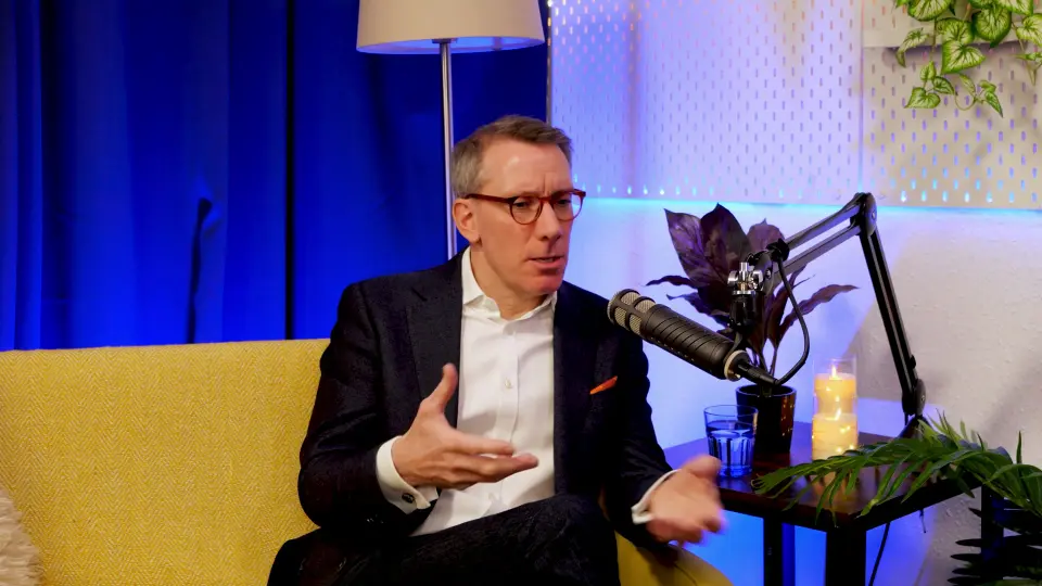 Geoff White gesturing mid-conversation on the BEYOND set, navy blazer, white shirt, podcast microphone in frame