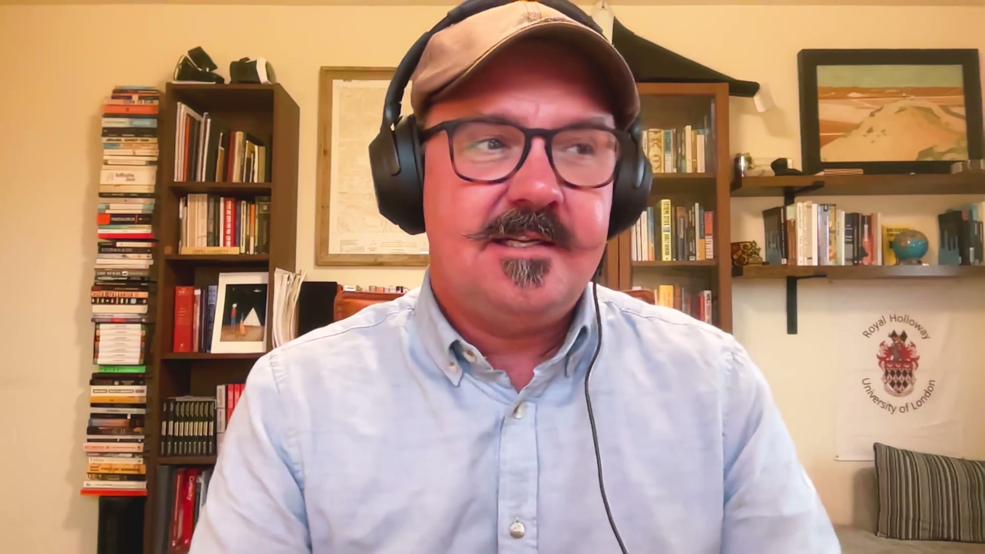 Bradley Garrett in headphones and cap smiling on The BEYOND Podcast, bookshelves and a Royal Holloway flag behind him