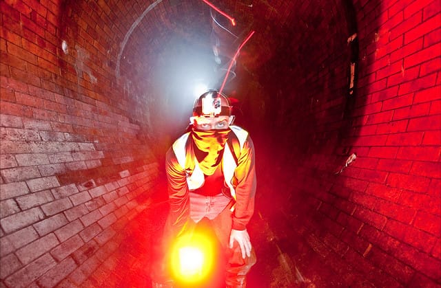 Bradley Garrett wading through the red-lit brick tunnel of the Westbourne sewer beneath London, headtorch and lantern blazing