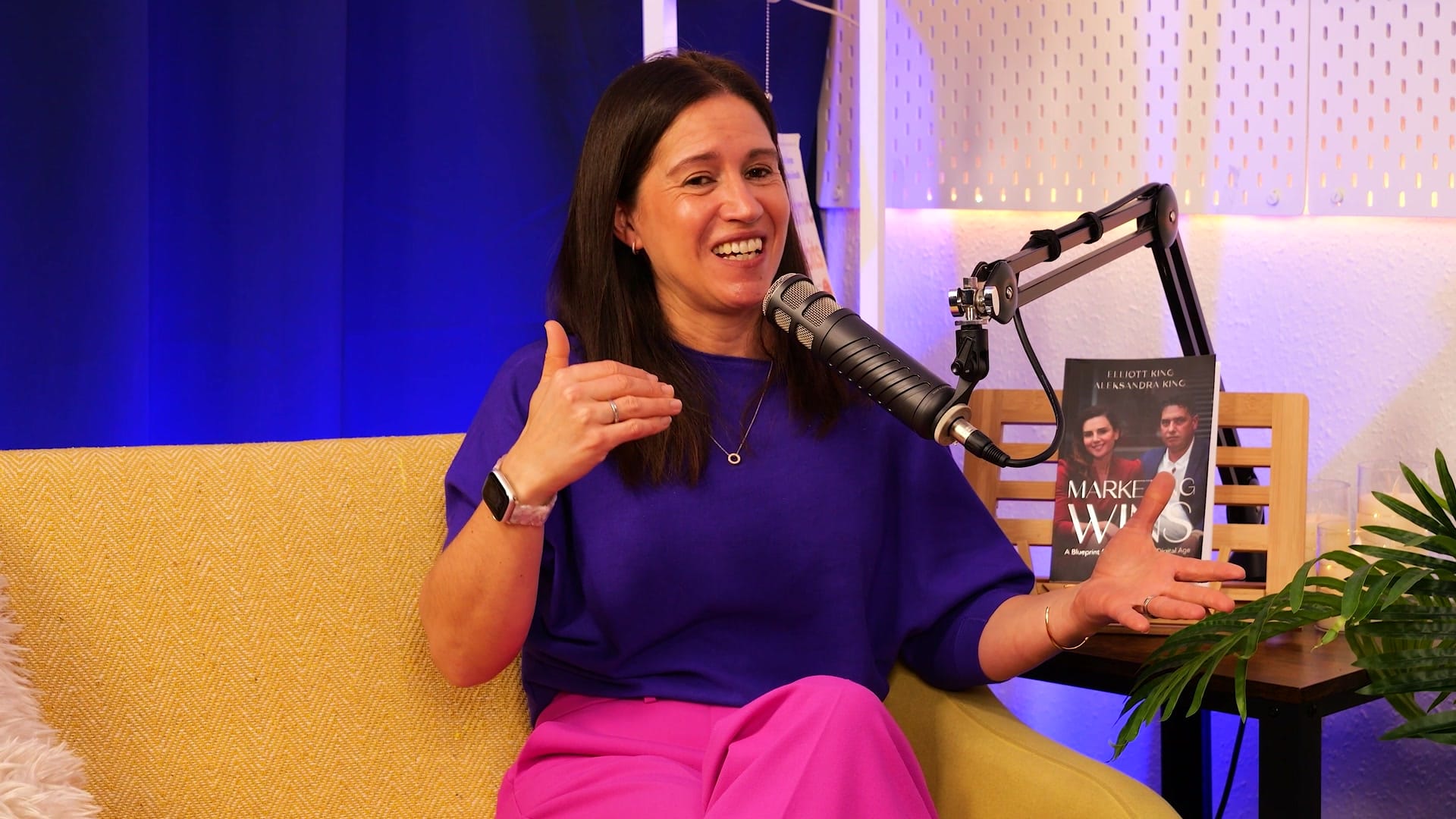 Anna Hemmings smiling mid-answer on the yellow sofa of The BEYOND Podcast set, in a purple top and pink trousers