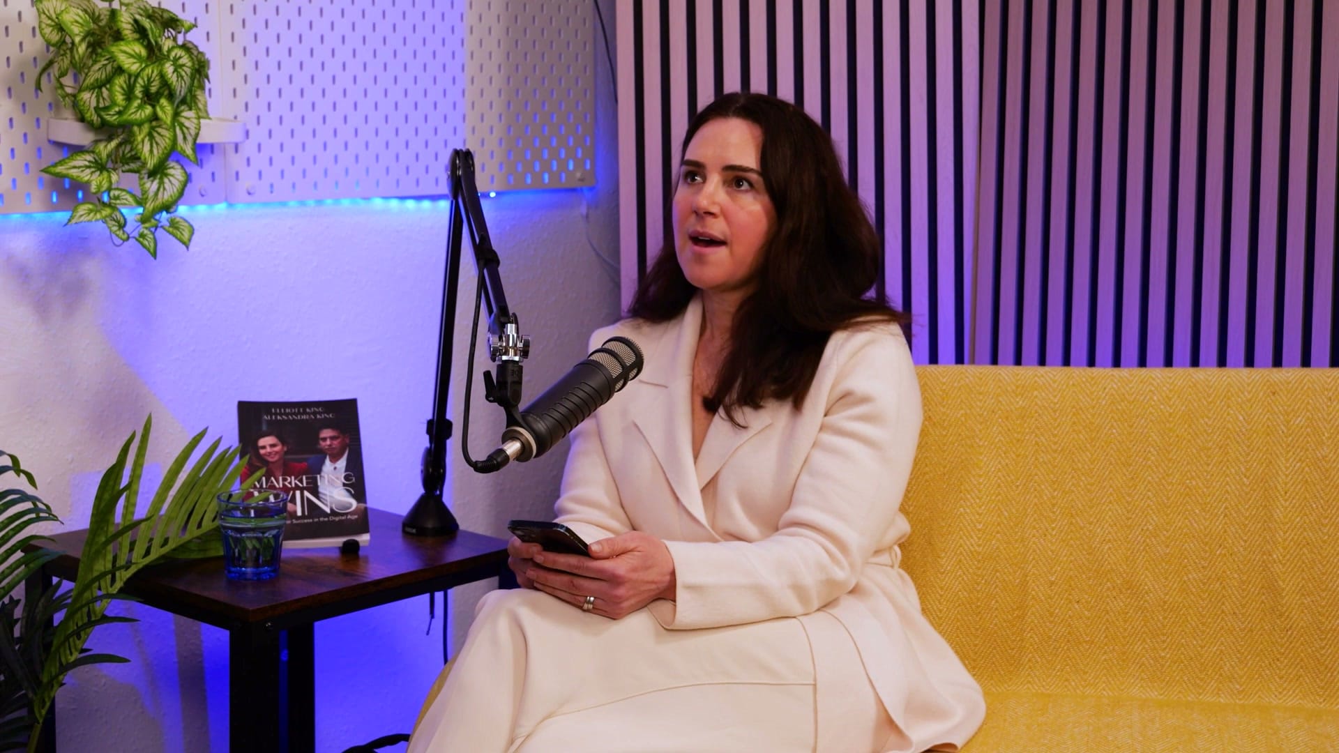 Aleksandra King in a cream suit, speaking into a microphone on a yellow sofa and holding her phone