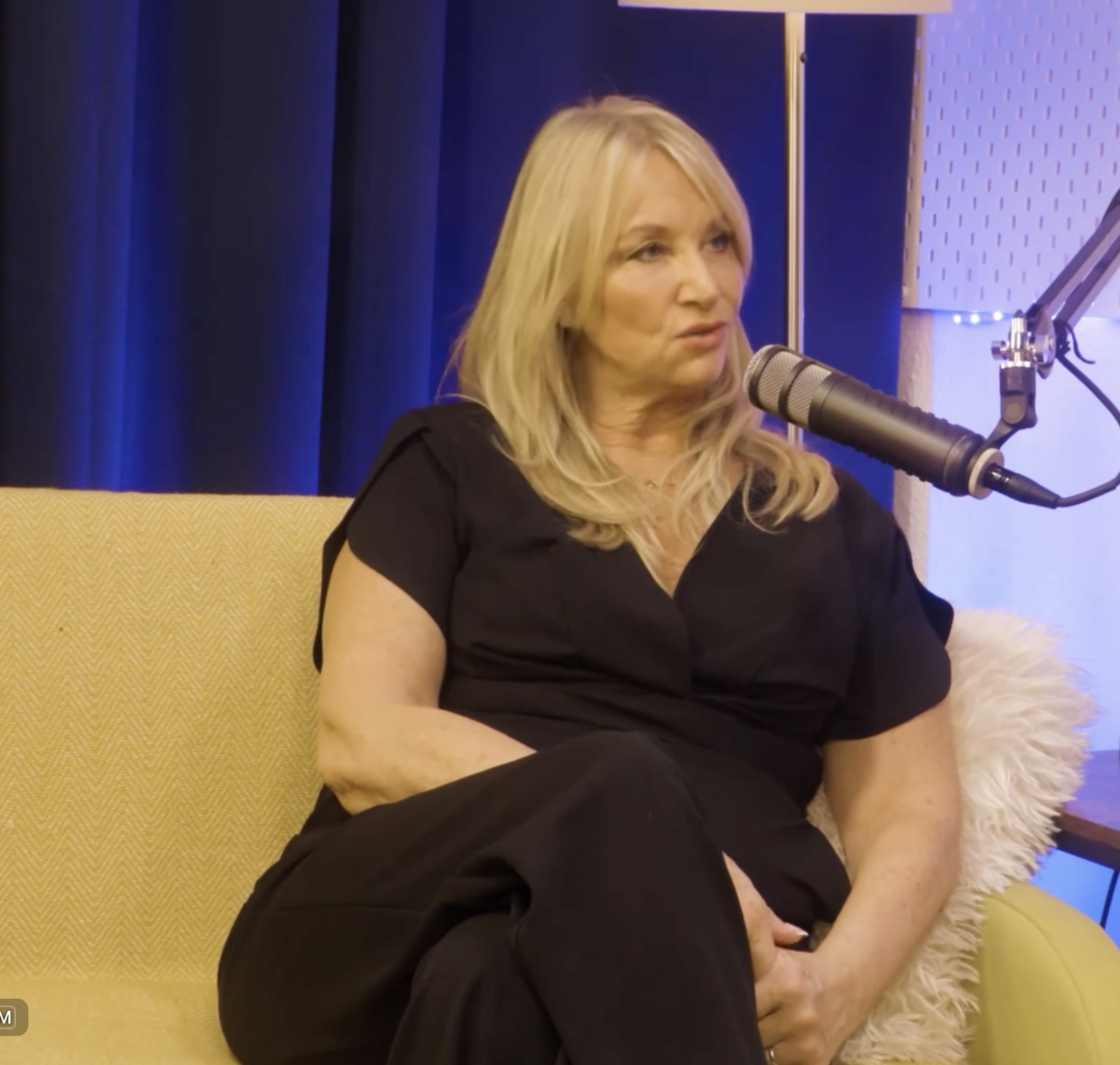 Deborah Mitchell speaking at the microphone during her BEYOND interview, seated on a yellow sofa in a black outfit