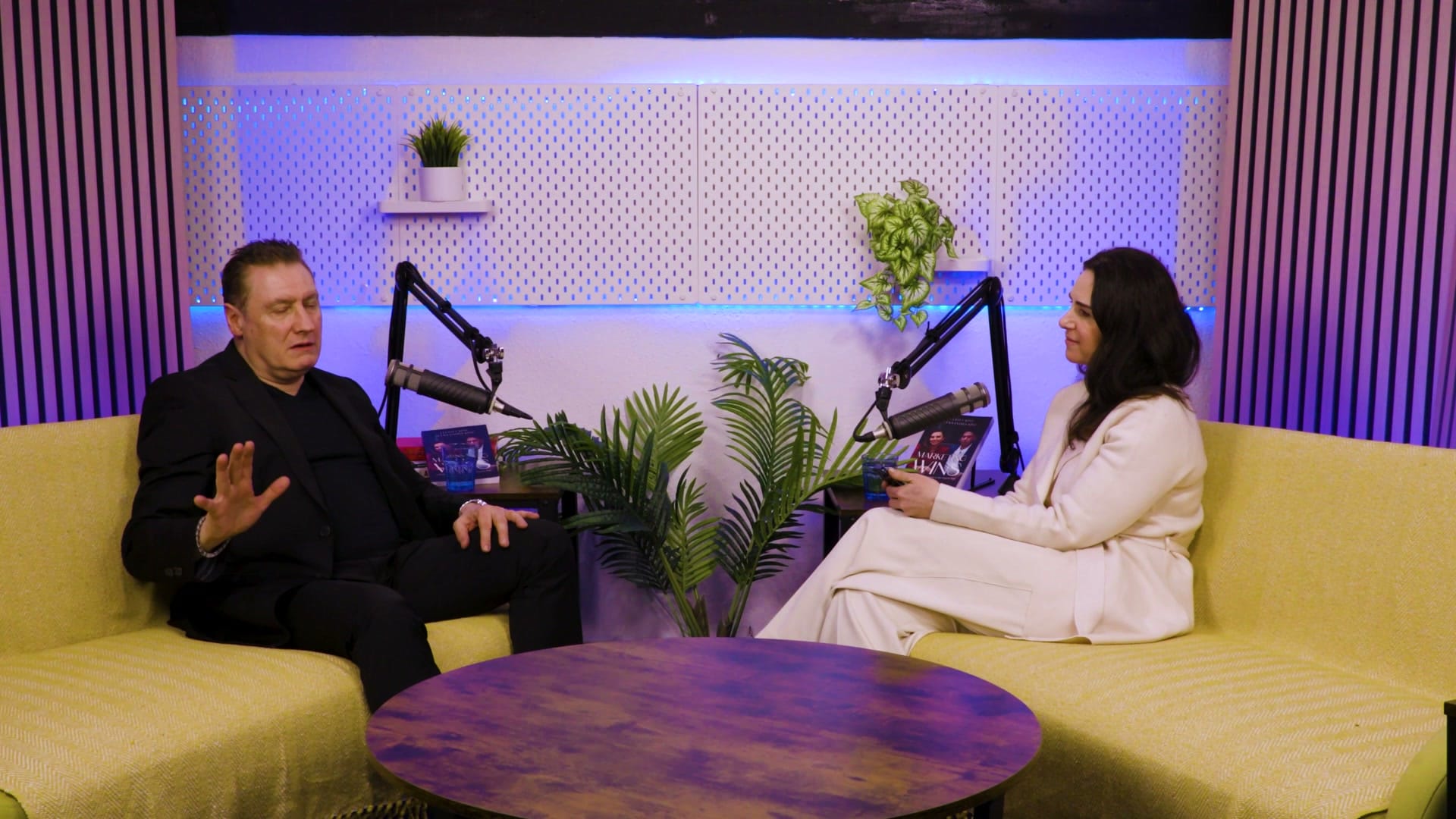 Grant Murrell and Aleksandra King seated on yellow sofas either side of a round table, microphones between them