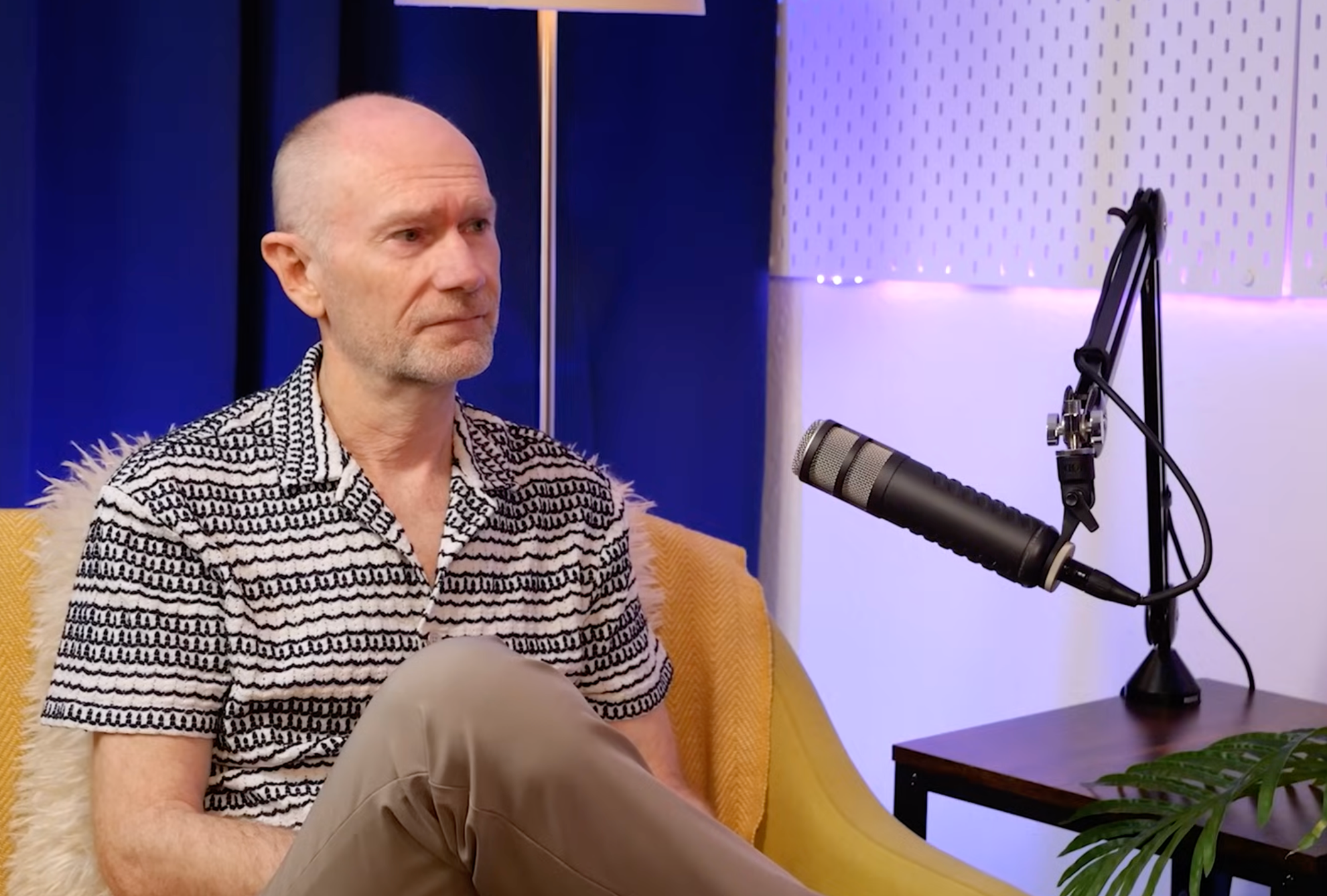 Robin Deller in a patterned shirt during his BEYOND interview, seated on a yellow sofa beside a boom-mounted microphone