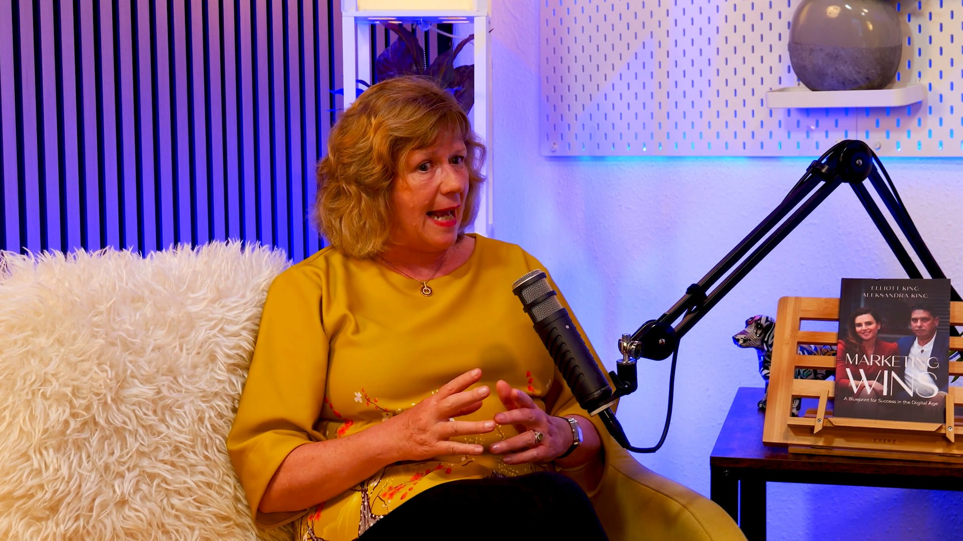 Sue Stockdale mid-sentence beside a studio microphone, hands clasped, on The BEYOND Podcast set
