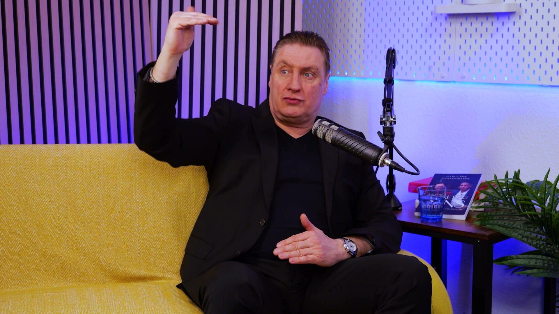 Grant Murrell raising one hand high above his head to illustrate a point, seated at a studio microphone