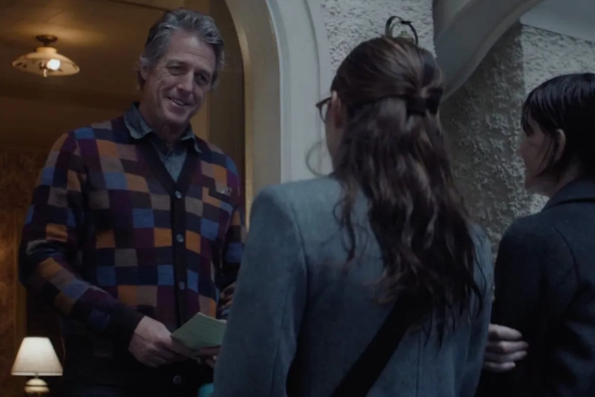 Screenshot from Heretic. Mr. Reed stands in his doorway in a color block cardigan, smiling at Sisters Paxton and Barnes.
