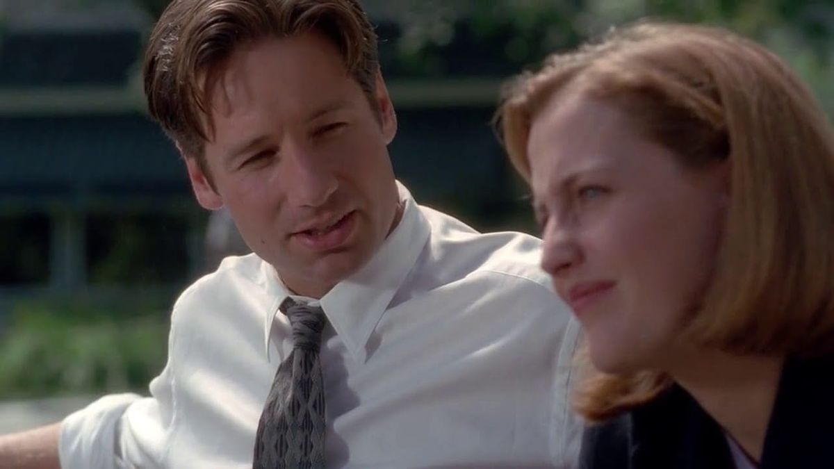 Mulder and Scully from The X-Files sitting next to each other on a bench. Mulder is saying something to her, and Scully is staring into the middle distance with a skeptical squint.