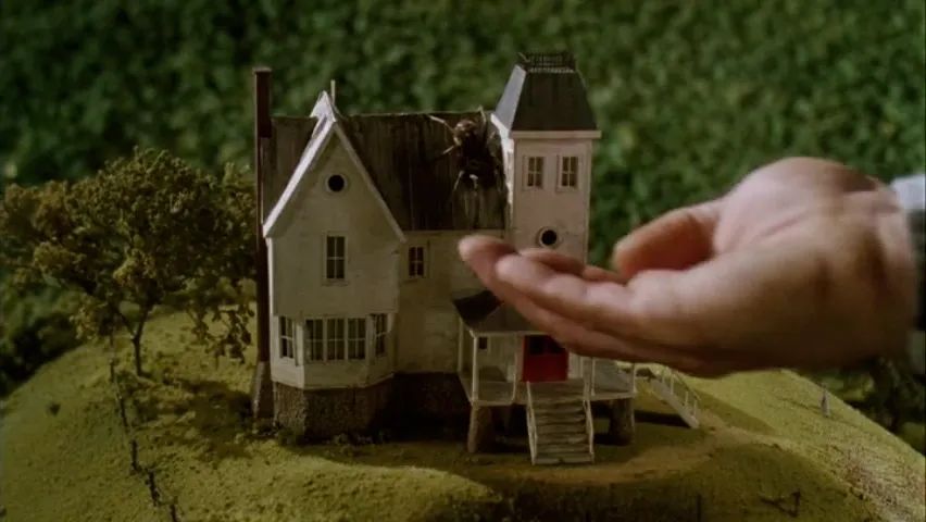 From Beetlejuice: A hand outstretched in front of a scale model of a house. A large spider crawls on the model's roof towards the hand.