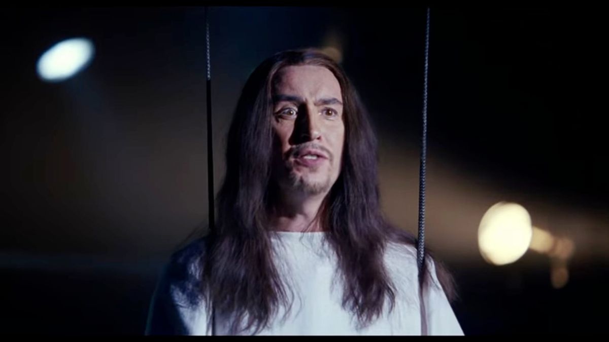 Steve Coogan with long hair and a white robe suspended from wires above a high school theater stage (playing Jesus).