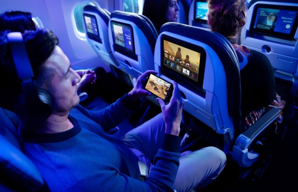 A man on a plane watches a video on his phone while videos play on the seat backs in front of him.