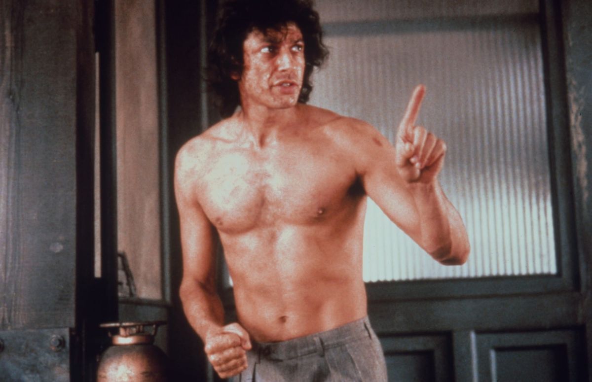 A shirtless Jeff Goldblum holds up his finger while talking to someone in The Fly.